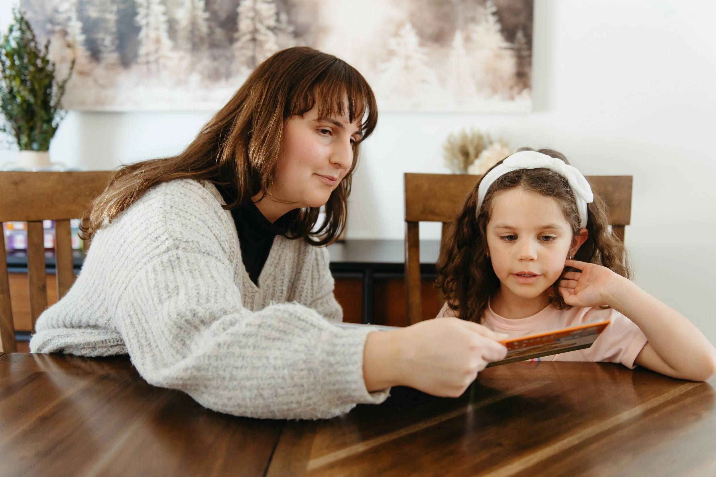 A woman and a young girl sitting at a wooden table, looking at a colorful book together indoors. The woman has brown hair and is wearing a cream sweater; the girl has curly brown hair with a white headband.