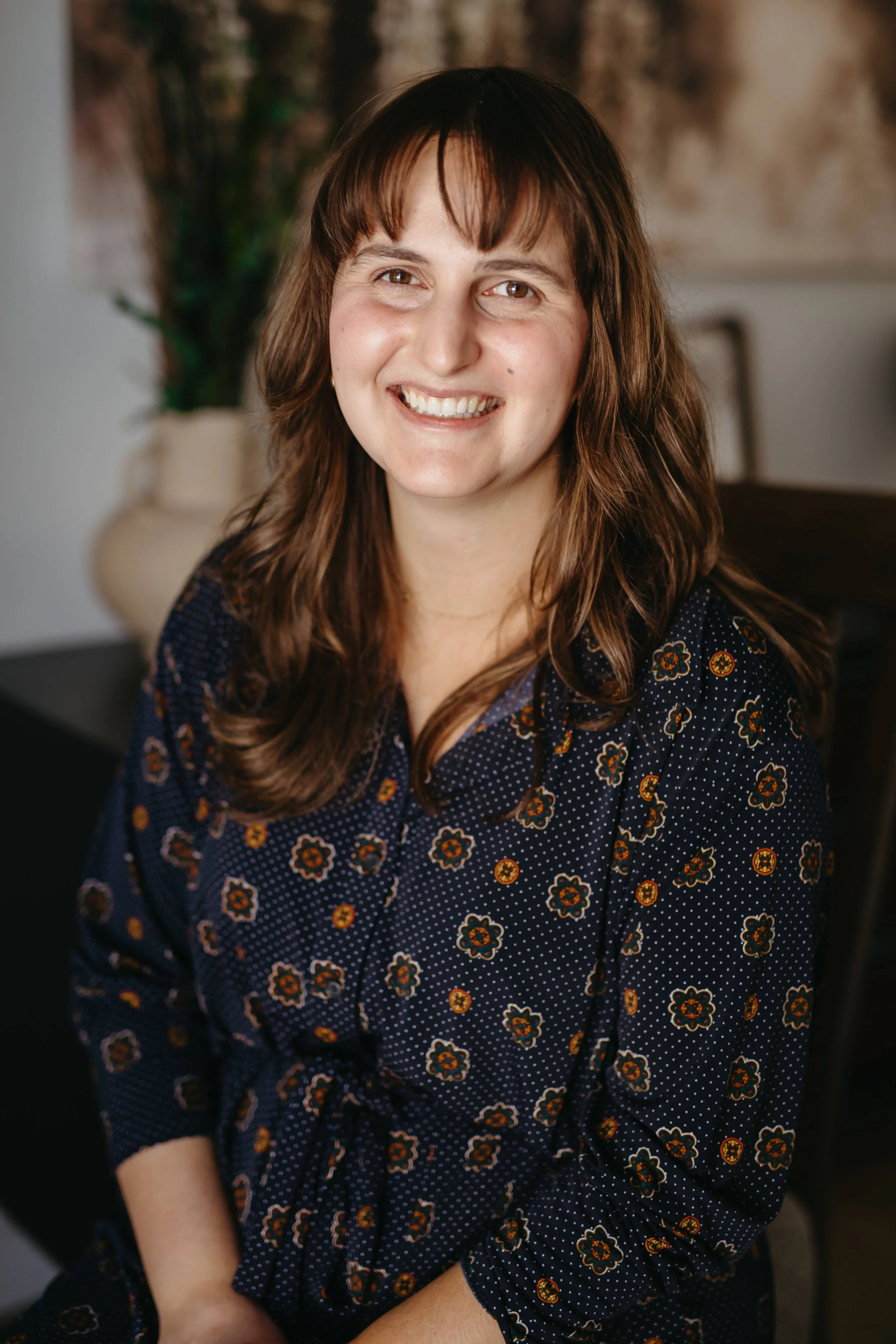 A woman (Halle Demchuk, founder of HAEPI SLP) with brown hair and bangs smiling, sitting indoors with a large vase and plant in the background, wearing a navy blue dress with a floral pattern.
