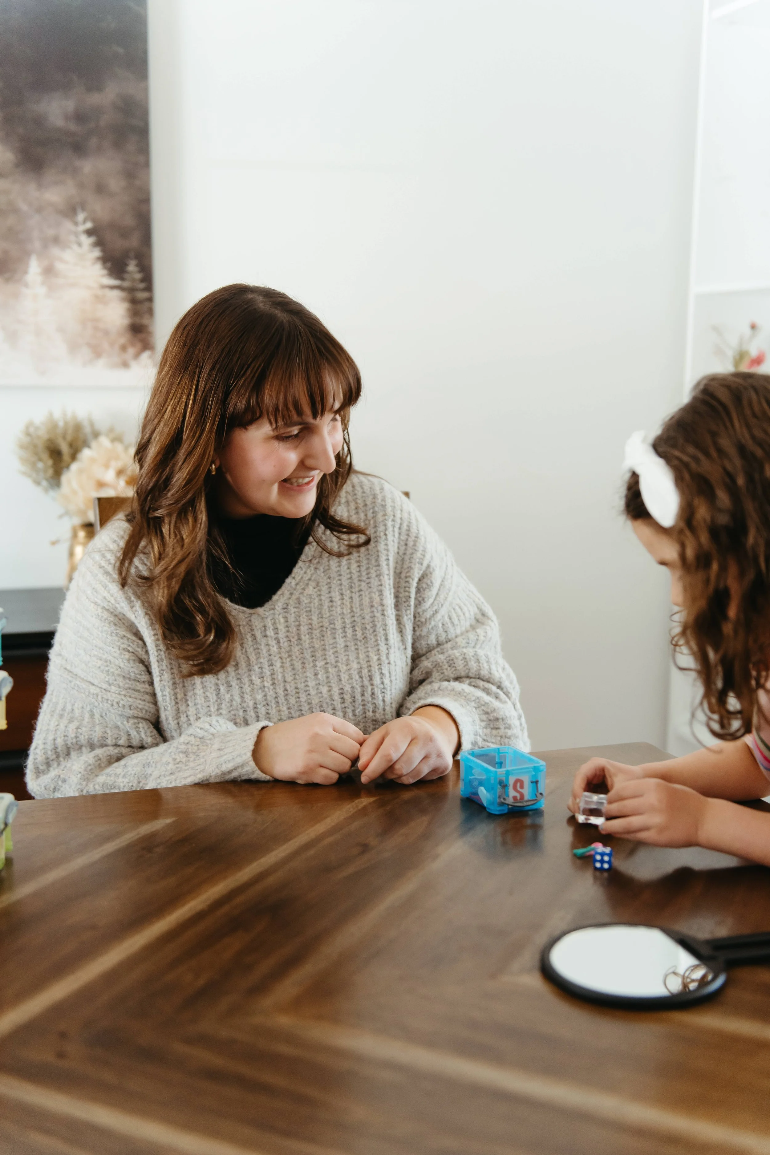 A woman and a young girl sitting at a wooden table, playing a game with small toys and dice, smiling at each other in a cozy, well-lit room.