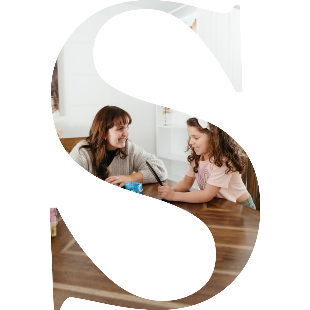 A woman and a girl are sitting at a wooden table, smiling and interacting with each other in a bright, cozy room.