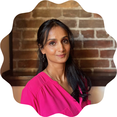 A woman (Asma Asad) with long black hair wearing a pink top standing in front of a brick wall.