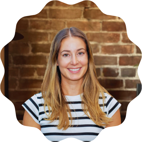 A woman (Akayla Olajos) with long blonde hair smiling, wearing a striped T-shirt, standing in front of a brick wall.