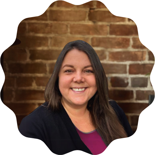 A woman (Ange Jewett) with long brown hair smiling at the camera, wearing a black blazer and a purple top, standing in front of a brick wall.
