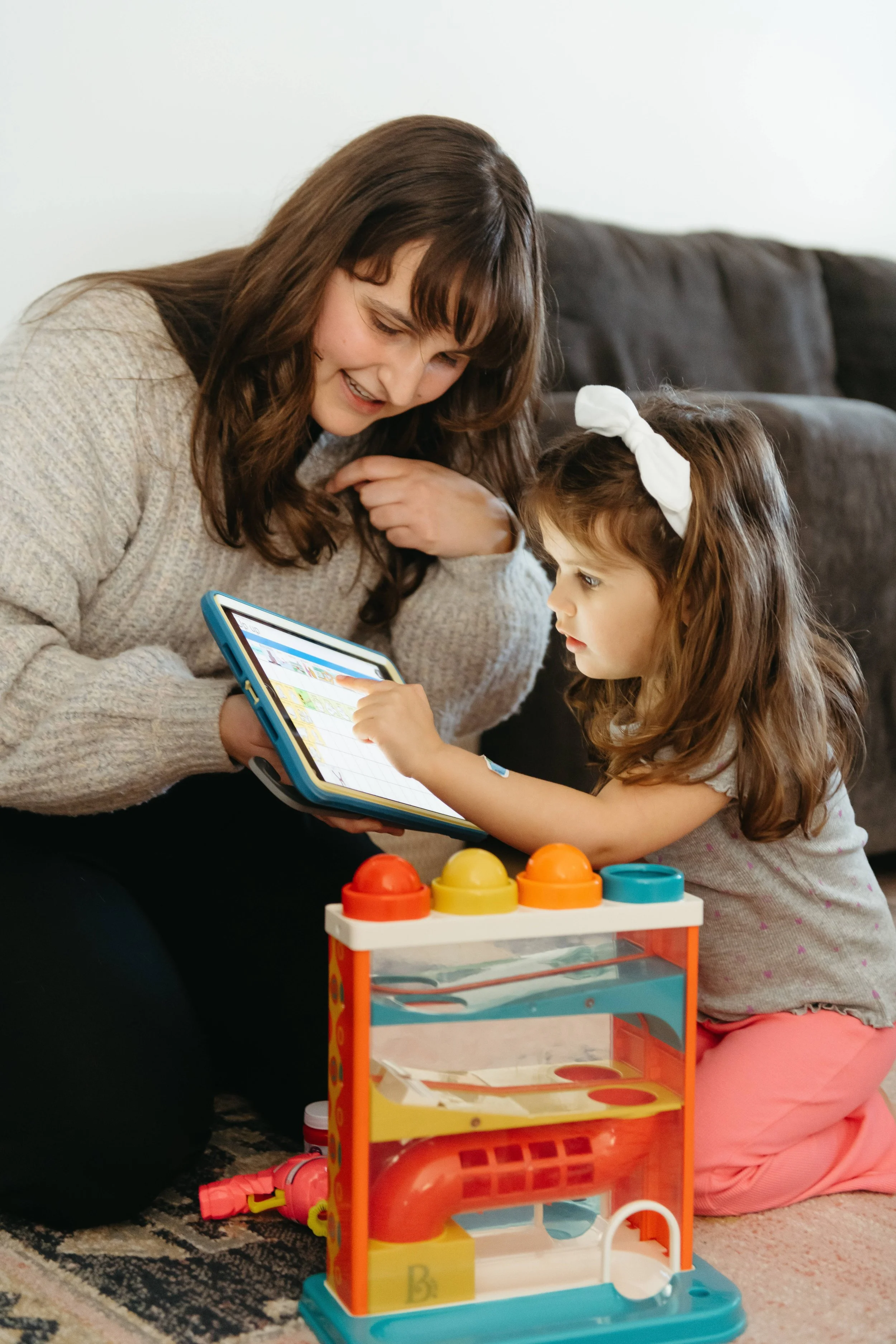A woman and a young girl playing with a tablet and a toy on a carpeted floor in a living room.