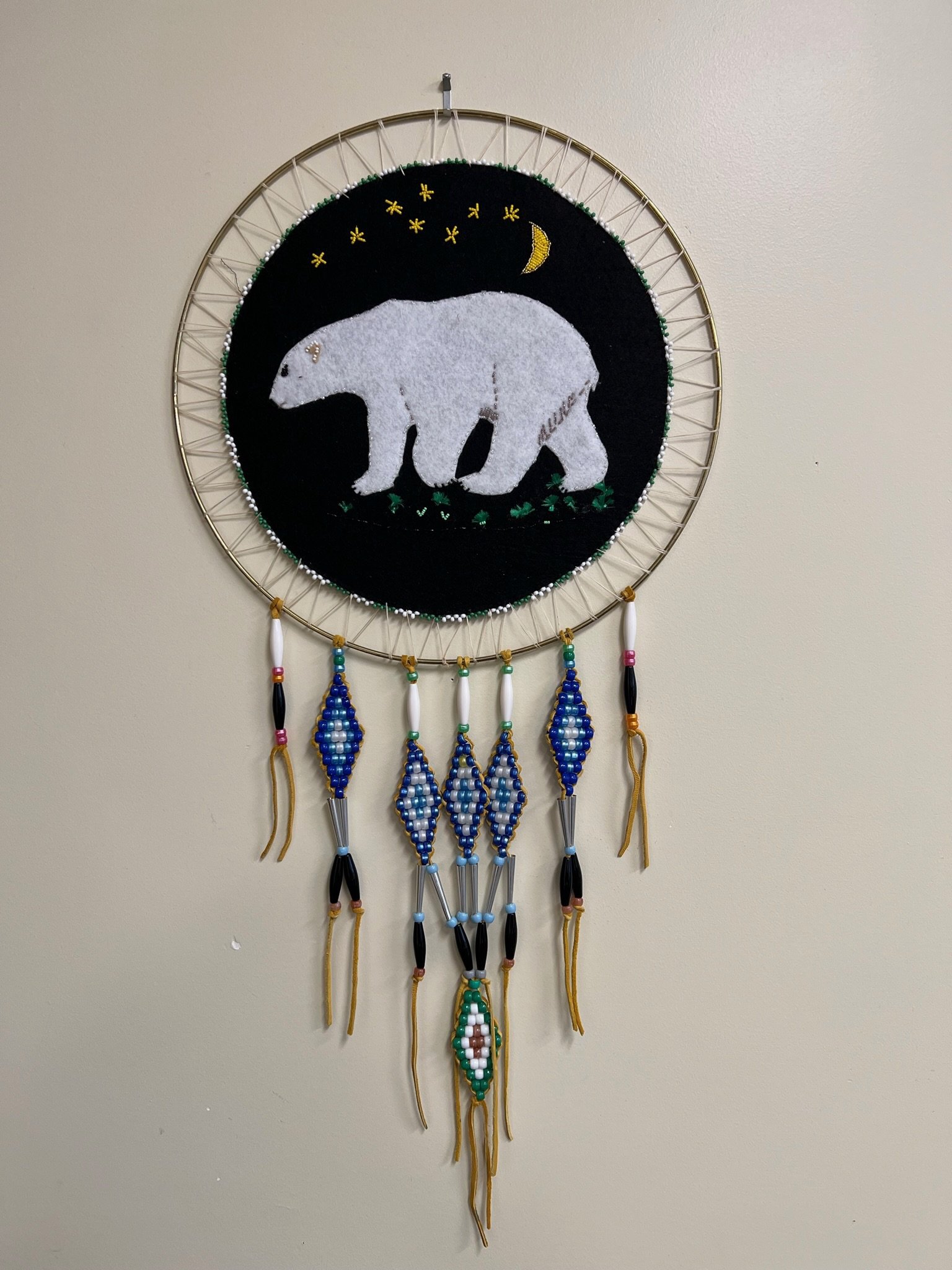A circular wall hanging of a bear walking on grass, surrounded by stars and a crescent moon, with beaded feathers hanging underneath.
