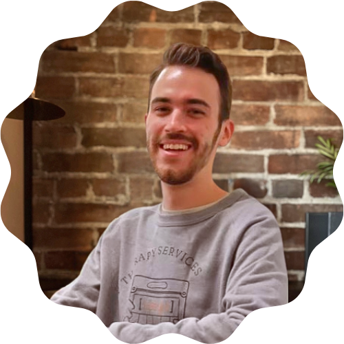 A smiling man (Zac McAlpine) with short dark hair and a beard wearing a gray sweatshirt, sitting in front of an exposed brick wall.