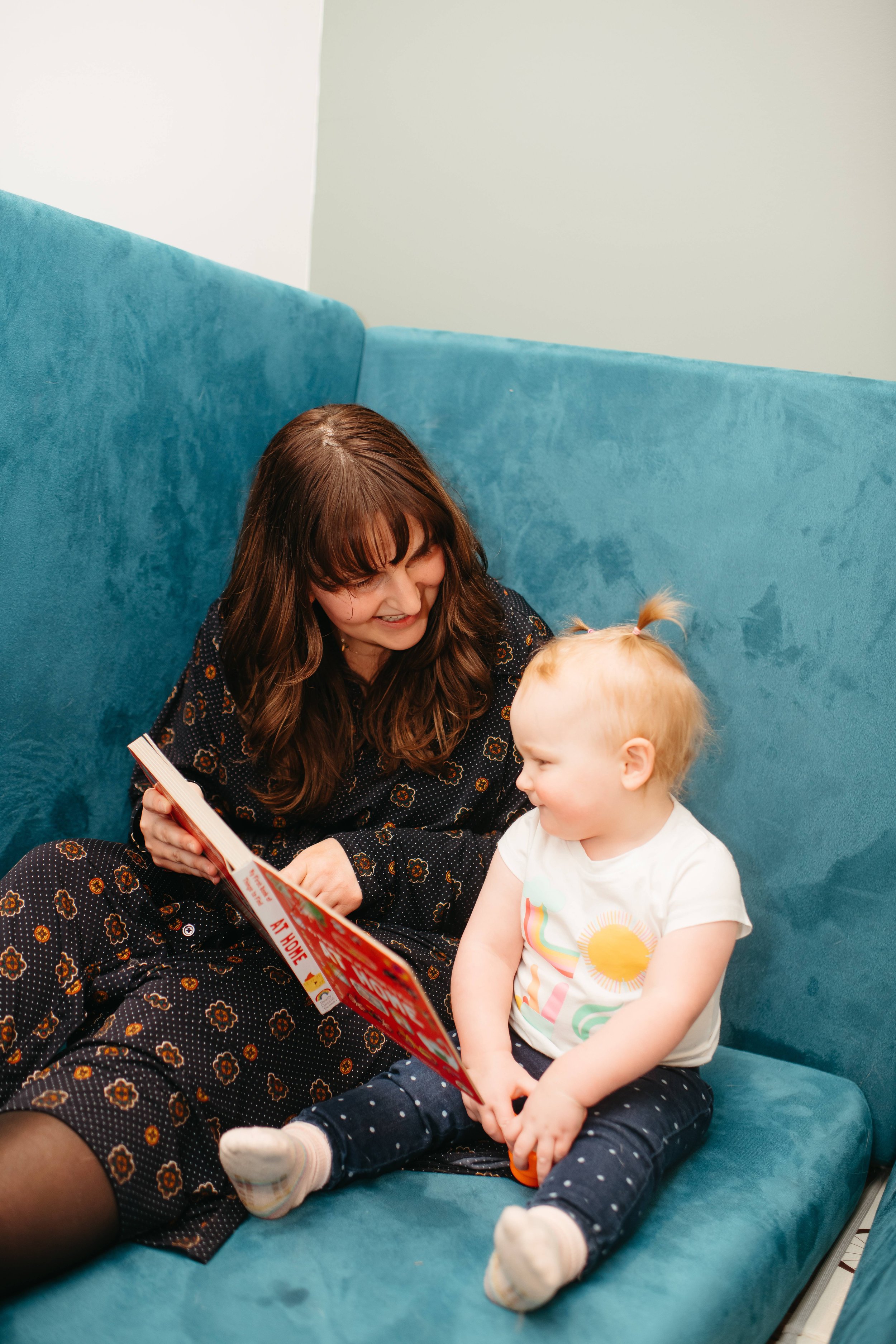 A woman and a young girl sitting on a blue couch, the woman reading a red book to the girl. The girl has blond hair in pigtails and is looking at the woman, who is smiling.