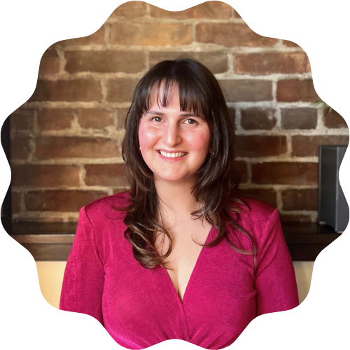 A woman (Halle Demchuk) with shoulder-length dark hair and a bright smile, wearing a pink top, standing in front of a brick wall.