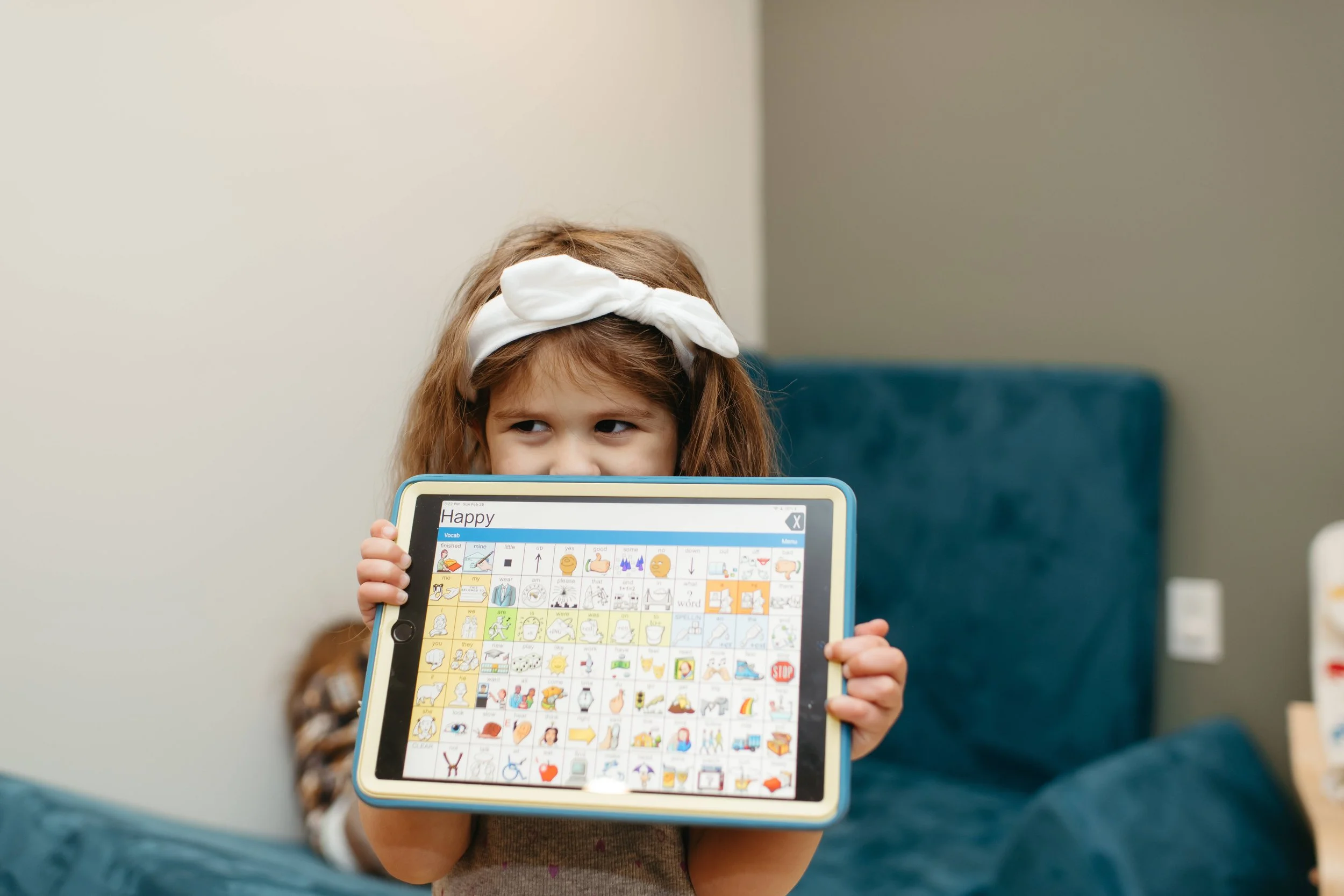 A young girl holding a tablet displaying a communication board with pictures and symbols, sitting on a teal couch.