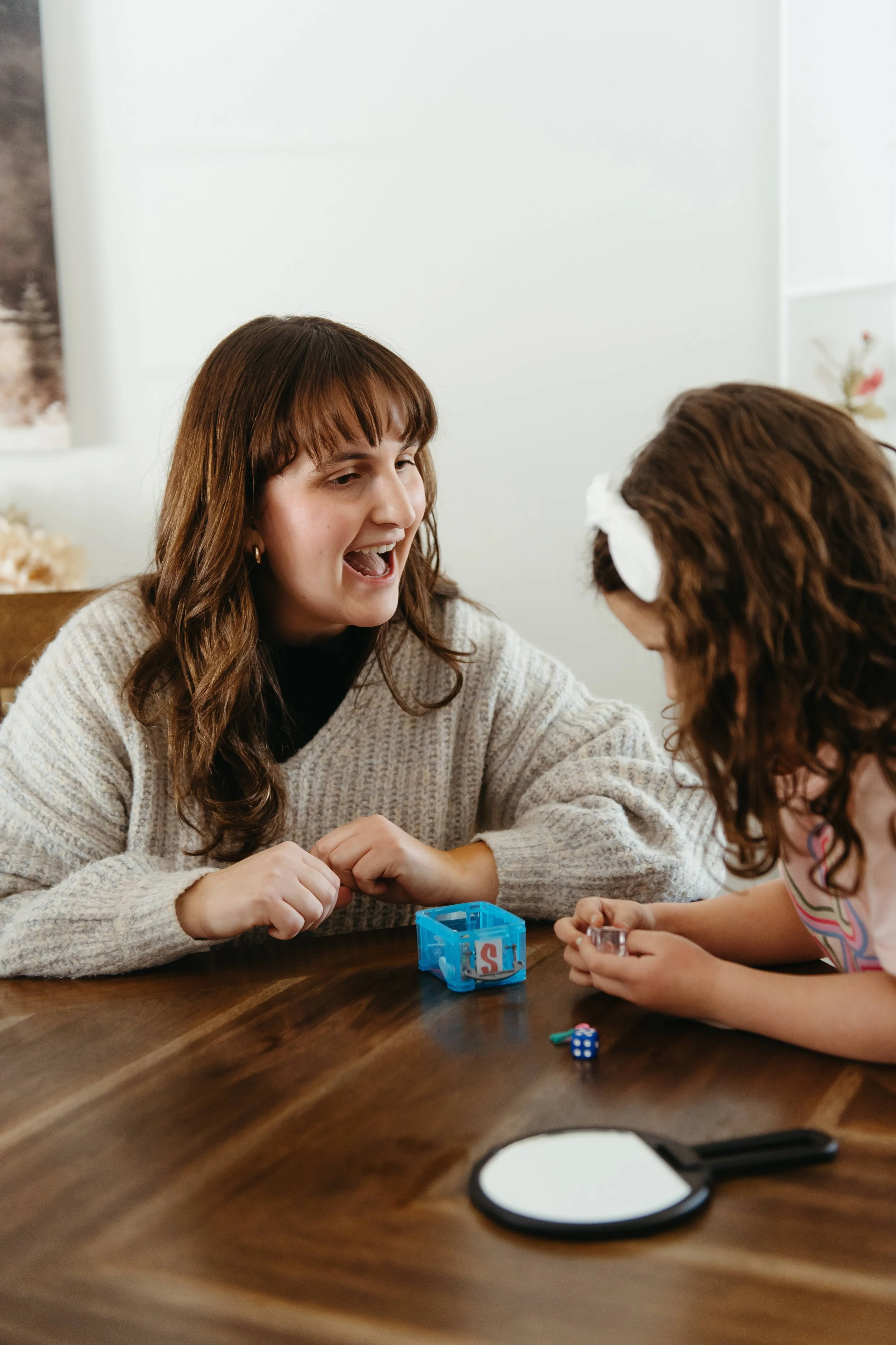 Two women playing a tabletop game with dice and cards in a cozy, well-lit room.