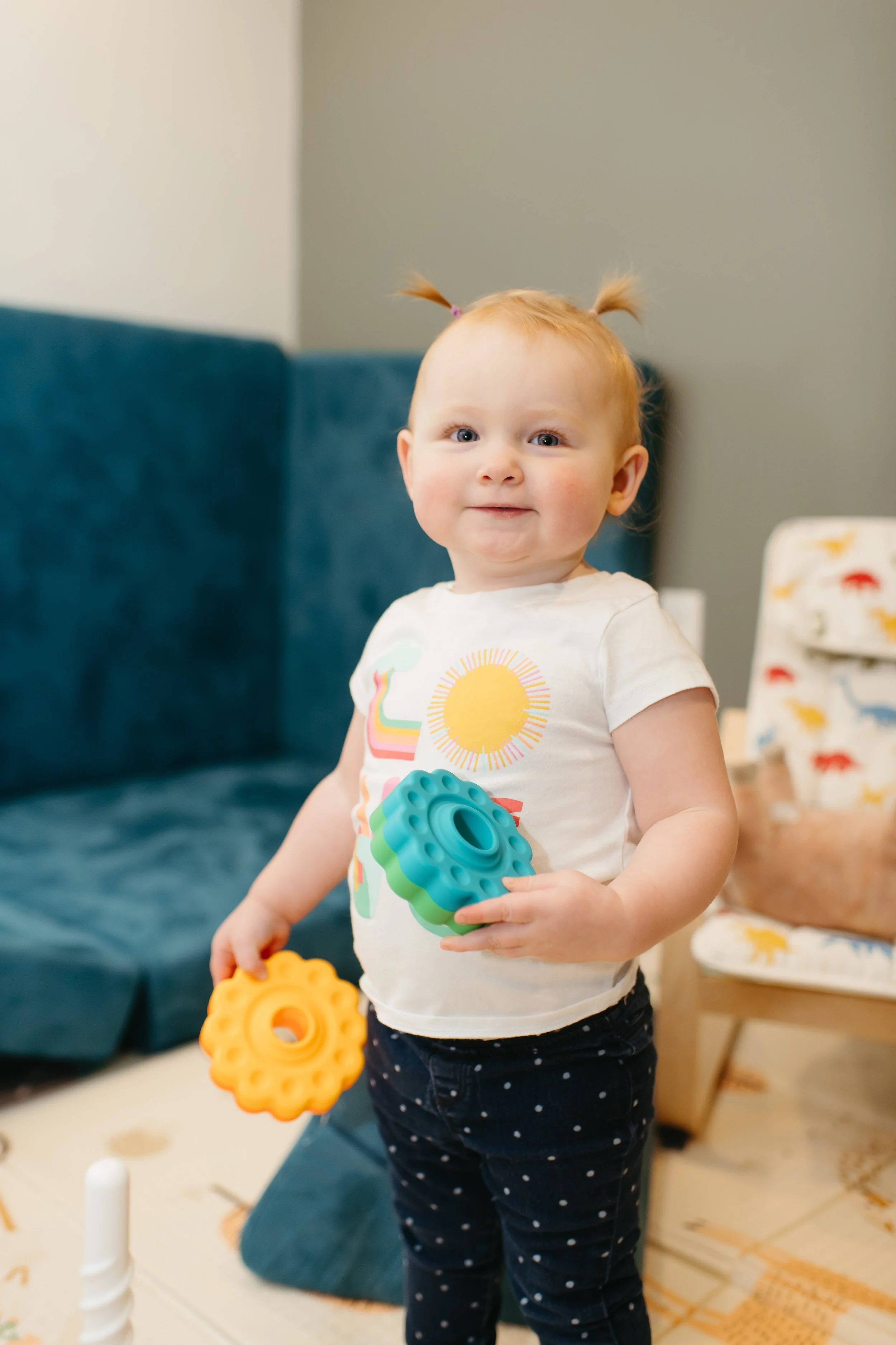 A young girl with red hair in pigtails holding colorful plastic toys inside a room with a blue couch and a chair with a animal print cover.