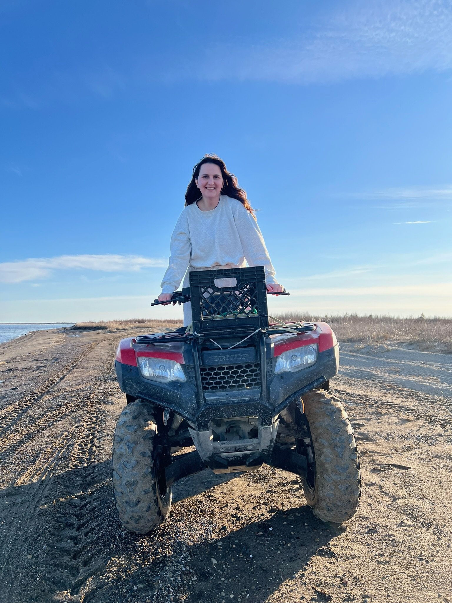 A woman smiling and riding a black and red all-terrain vehicle on a sandy terrain with a clear blue sky in the background.