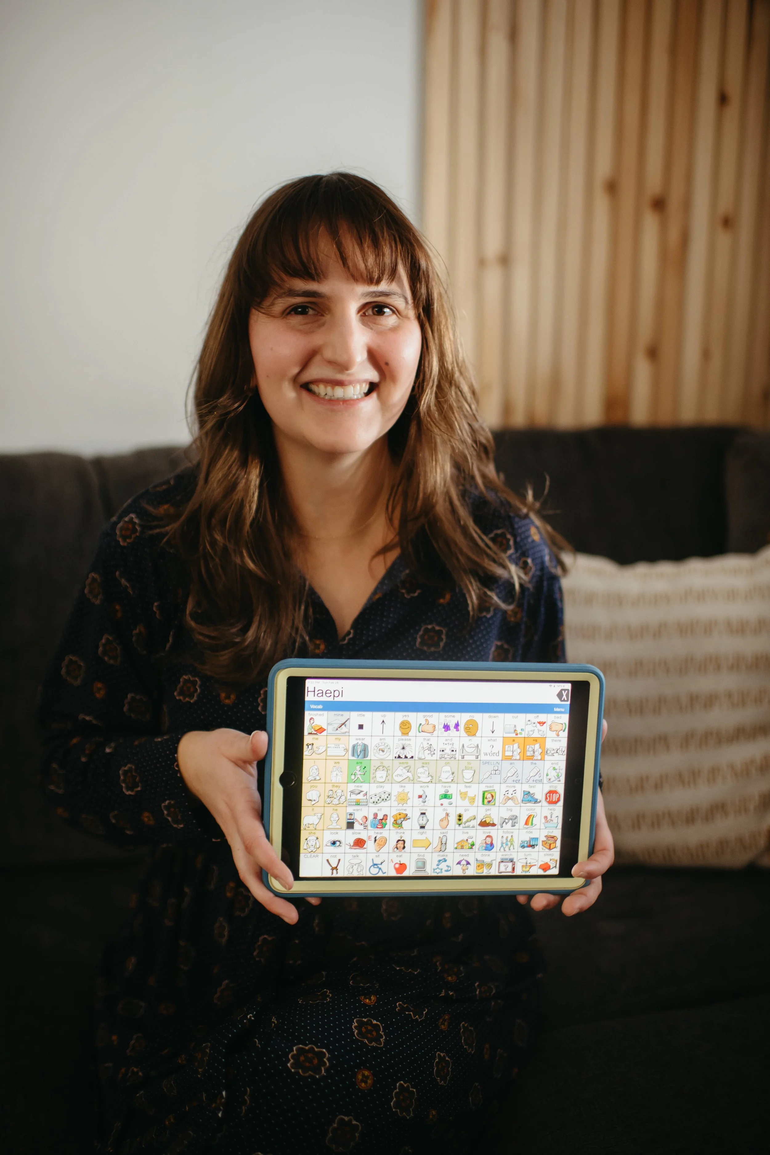 A smiling woman (Halle Demchuk, SLP) holding a tablet with a picture communication board (AAC) displayed on the screen.