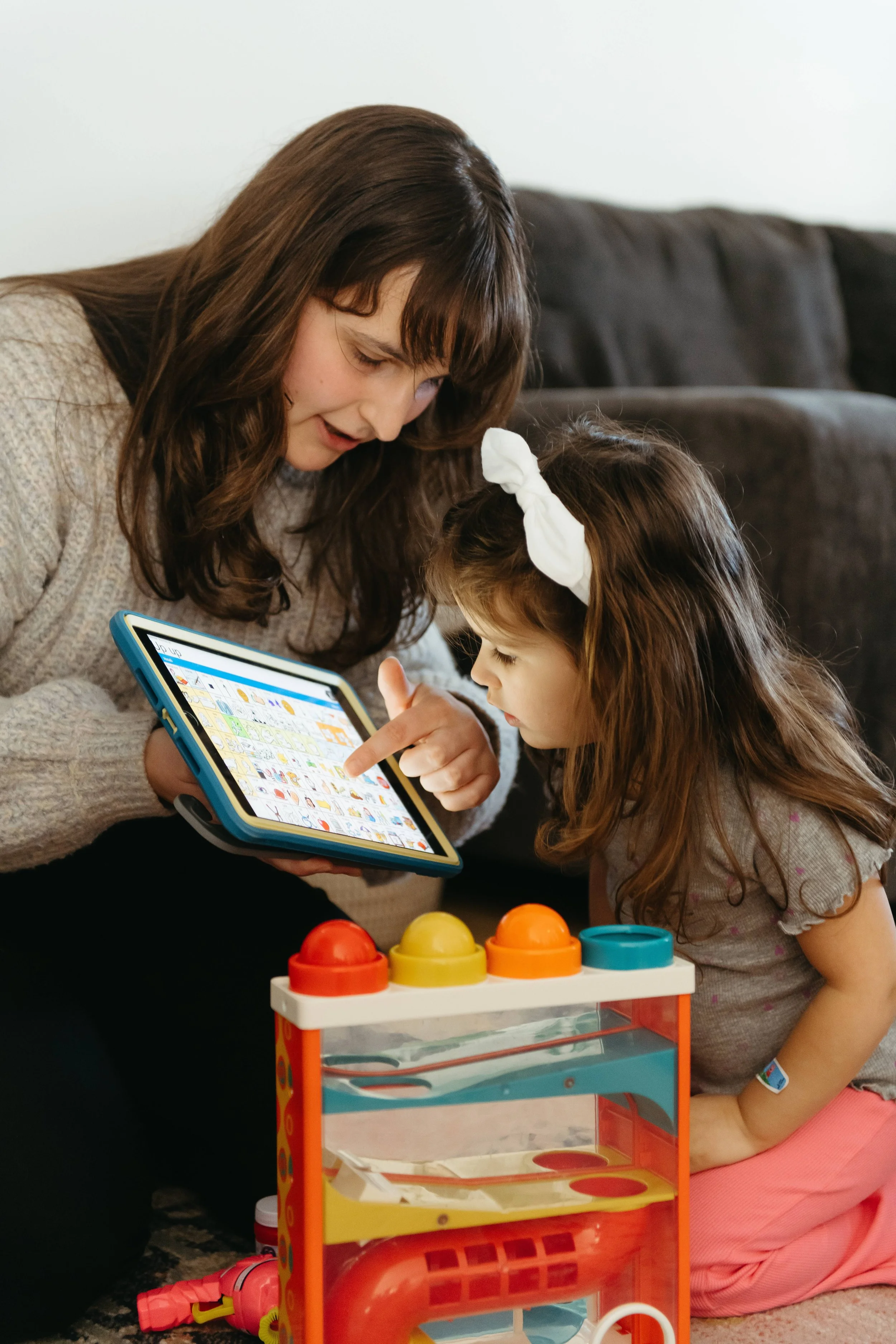 A woman and a young girl are sitting on a carpeted floor, looking at a tablet together. The girl is kneeling, and the woman is pointing at the tablet. There are colorful toys in front of them and a dark couch in the background.