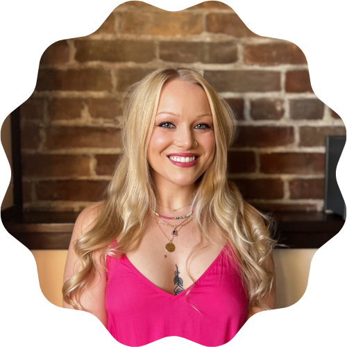 A smiling woman (Piper Rasmussen) with long blonde hair in a pink top standing in front of a brick wall