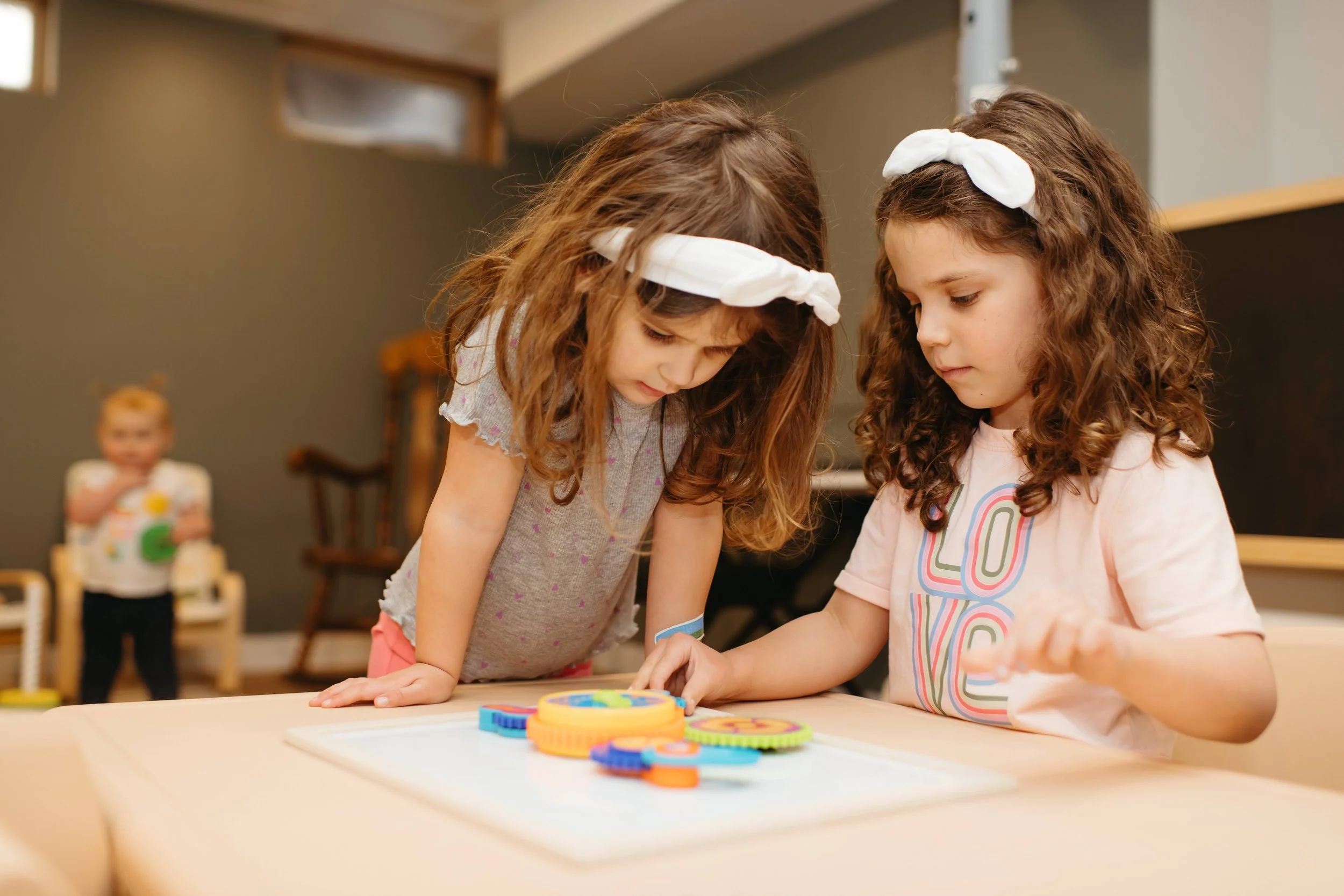 Two young girls with curly hair and white headbands playing a board game at a table, with a toddler observing in the background.