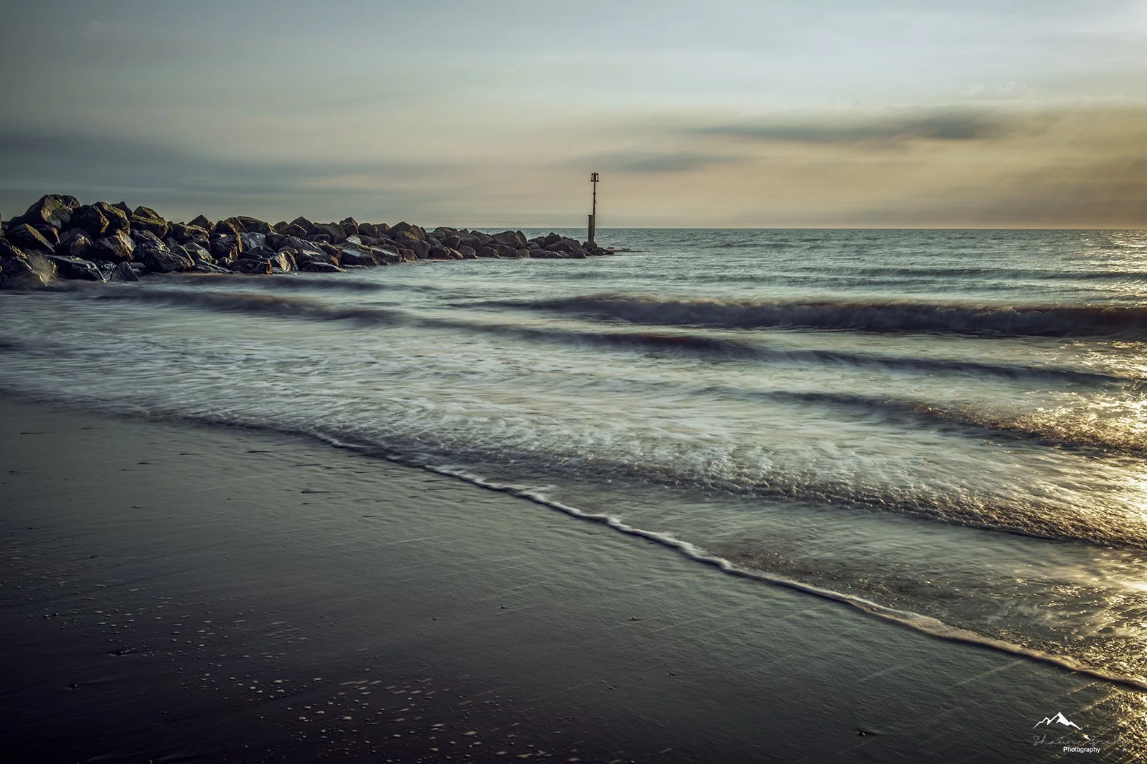 Sunrise over the North sea with waves gently crashing on the sandy beach and a rocky jetty extending into the water.