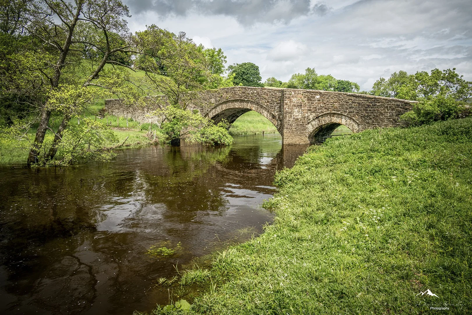 A rustic stone arch bridge over a calm river, surrounded by green trees and grass under a cloudy sky.
