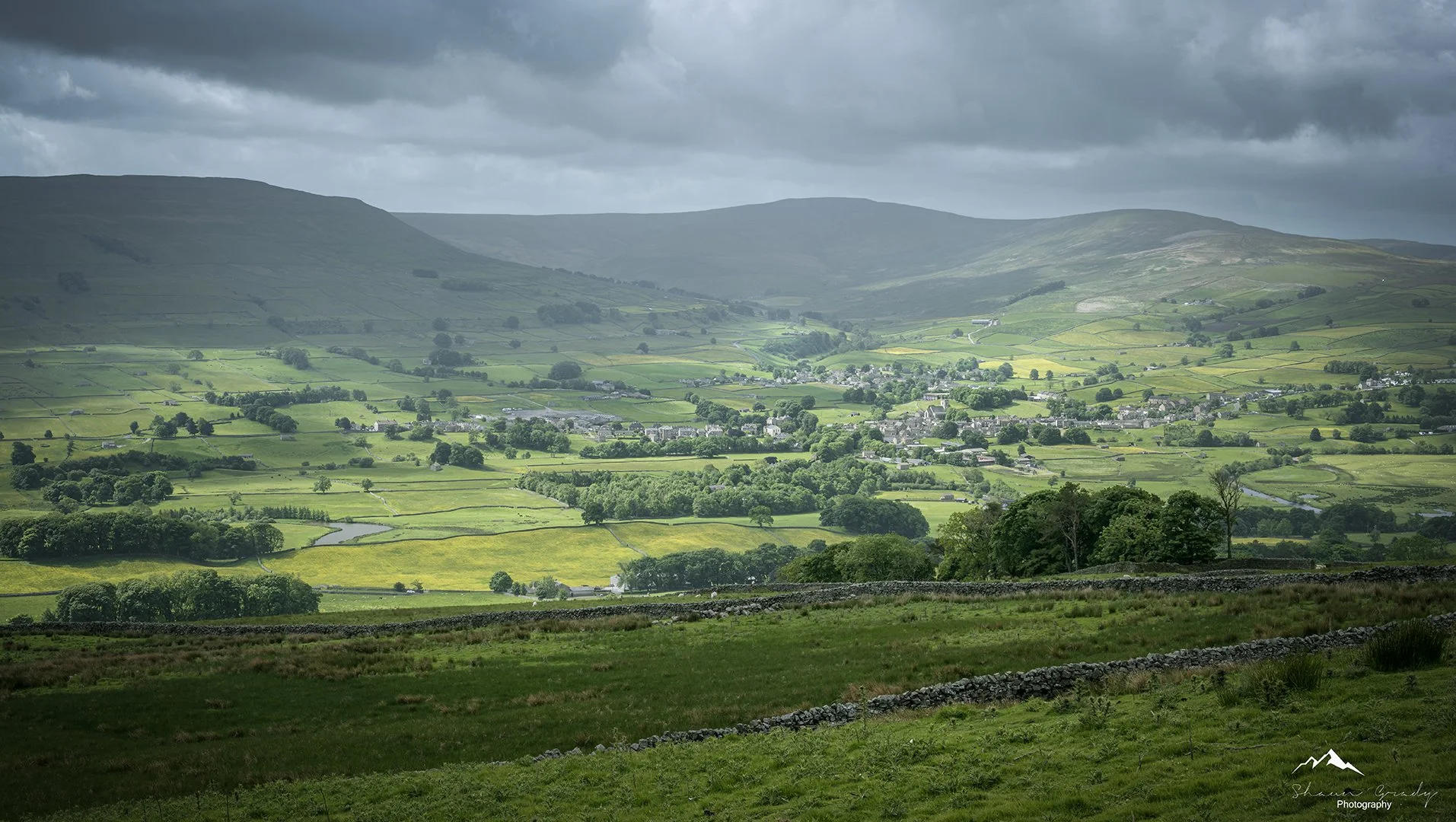 A lush green valley with scattered trees, fields separated by stone walls, a river, and a small village. Mountainous terrain under a cloudy sky in the distance.