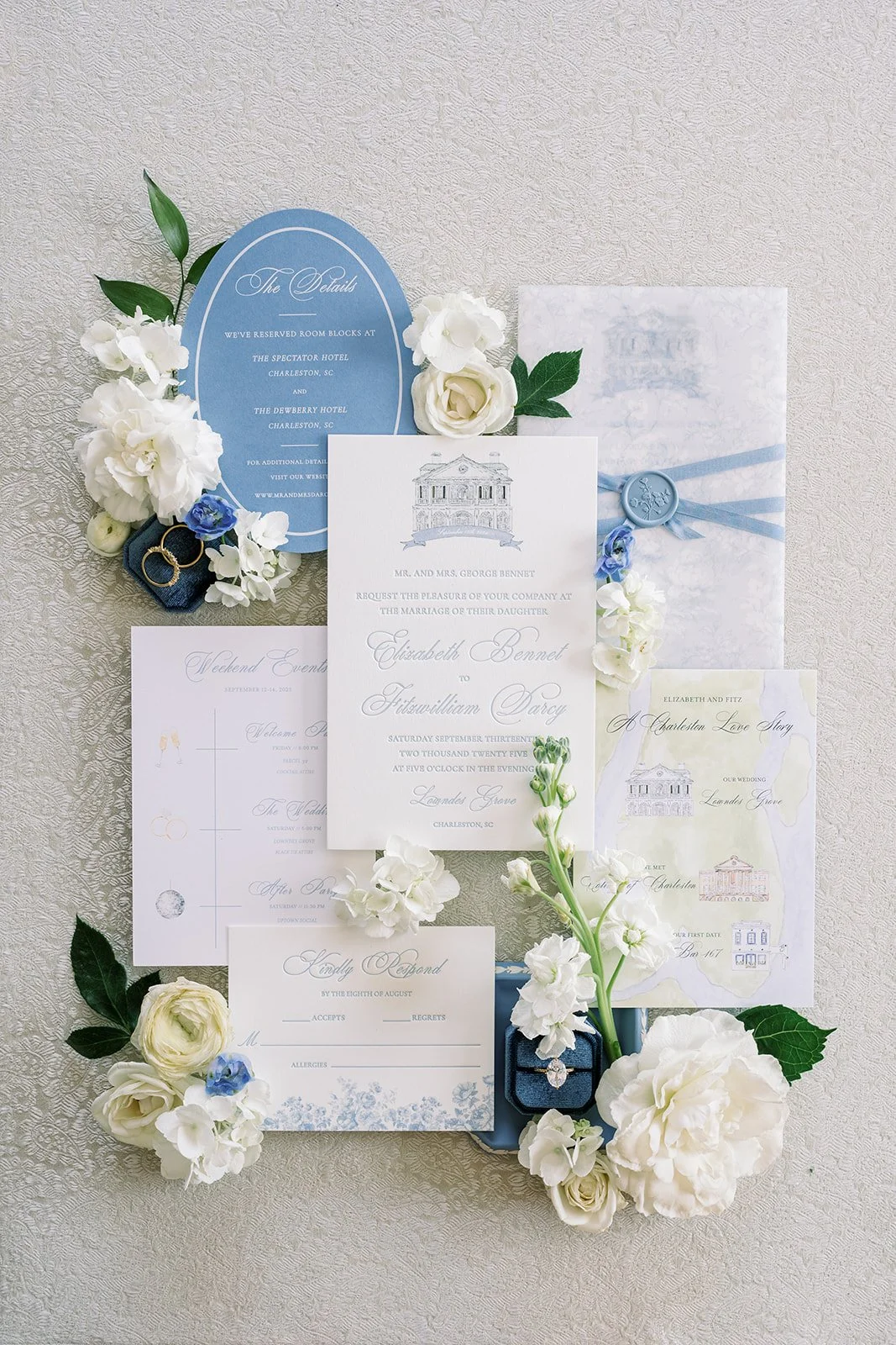 Wedding invitation suite with white and blue accents, surrounded by white flowers, green leaves, and wedding rings, on a textured white surface.