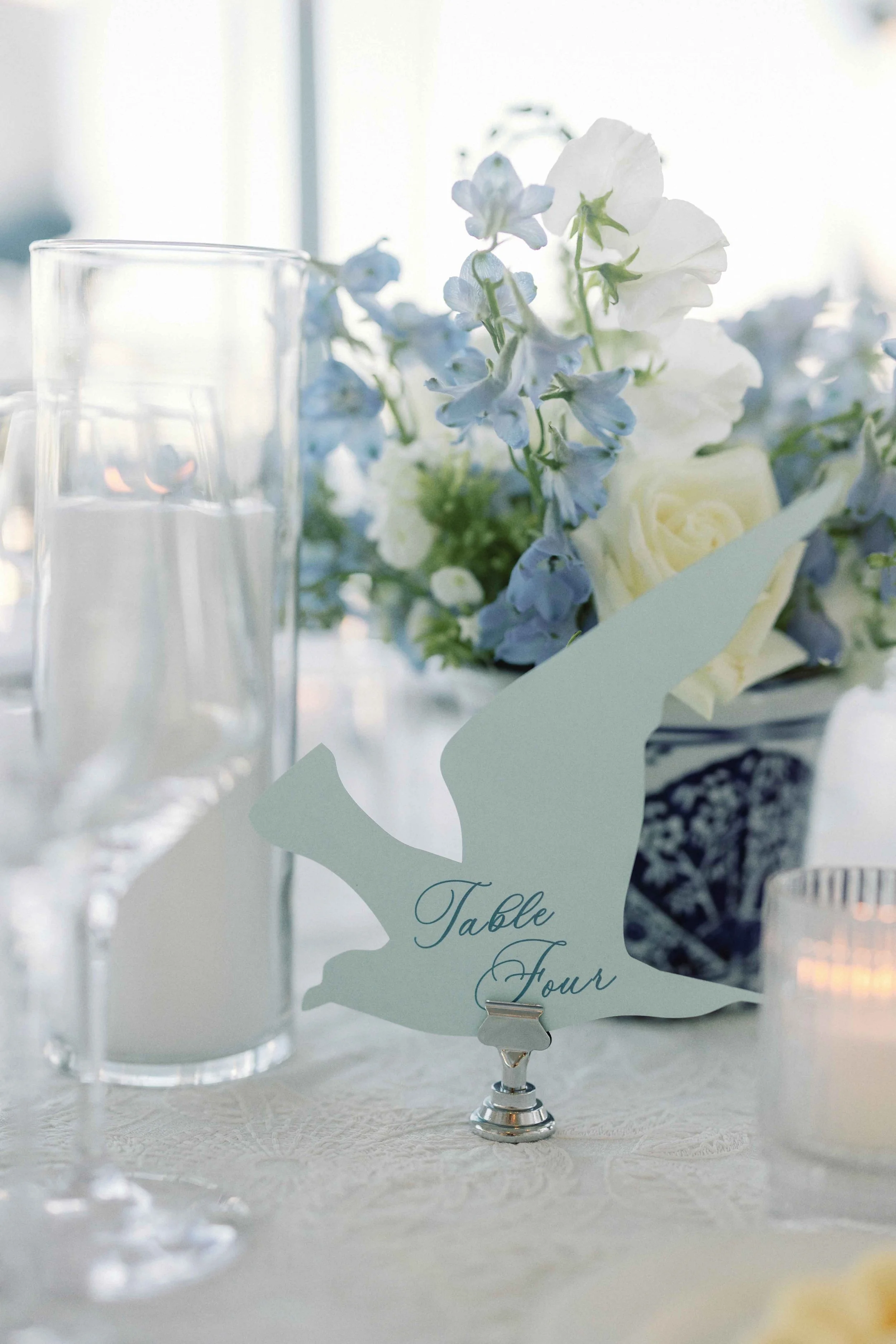 A bird-shaped cut-out featuring a table number on it next to glassware, florals, and candles