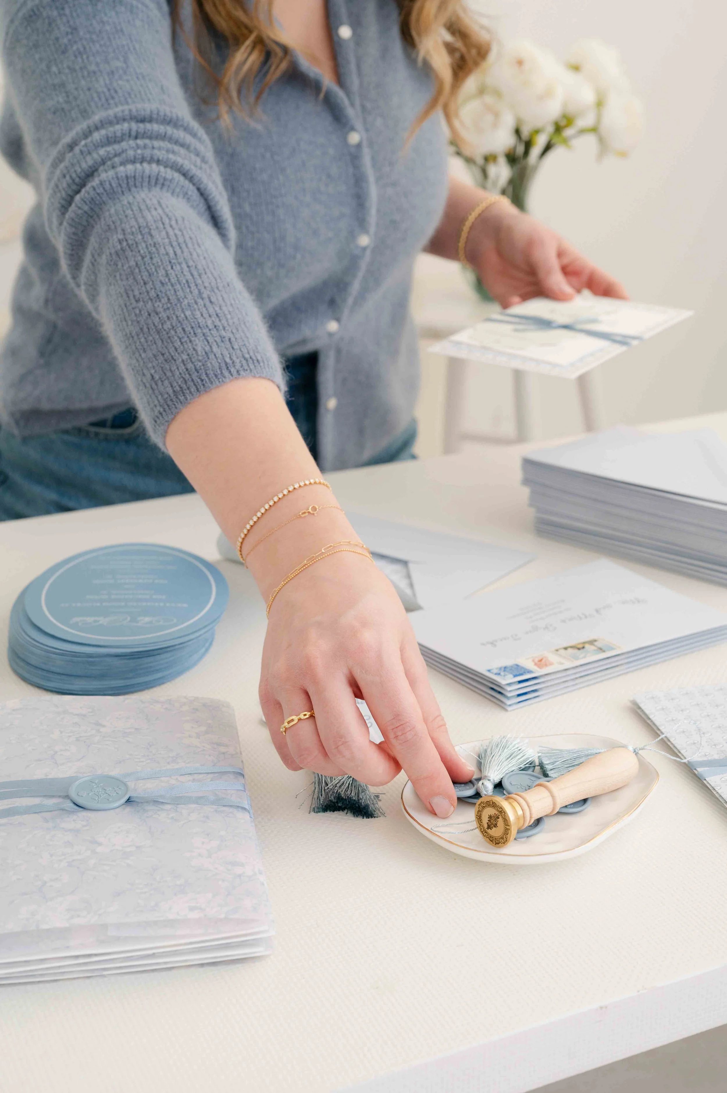 Woman wearing a blue sweater and jewelry, preparing wedding stationery with blue and white accents, on a white table with wedding invitations, envelopes, and stamps.