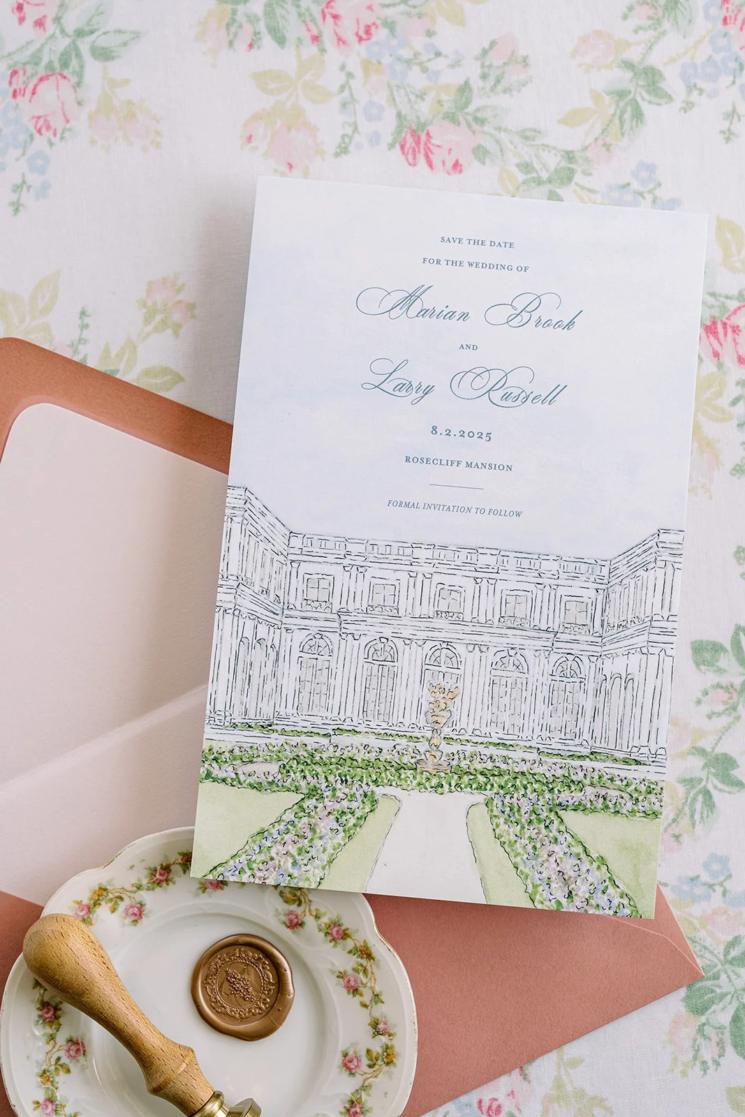 A wedding invitation card featuring a line drawing of a mansion, with floral accents, placed on a floral tablecloth, alongside a small dish with a wax seal and a sealing stamp.