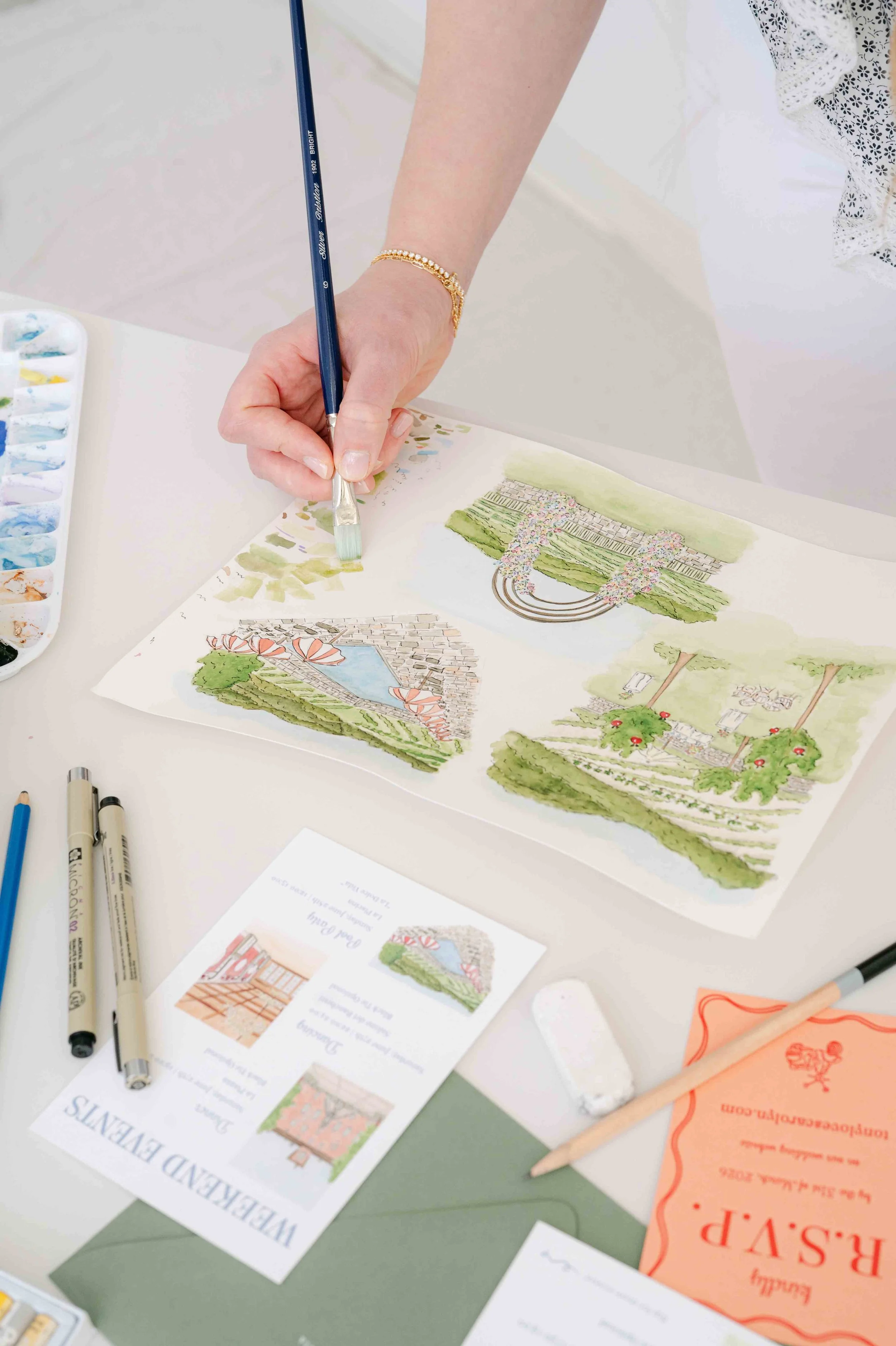 Person creating watercolor illustrations of outdoor landscape scenes on paper, surrounded by art supplies like pens and a paint palette.