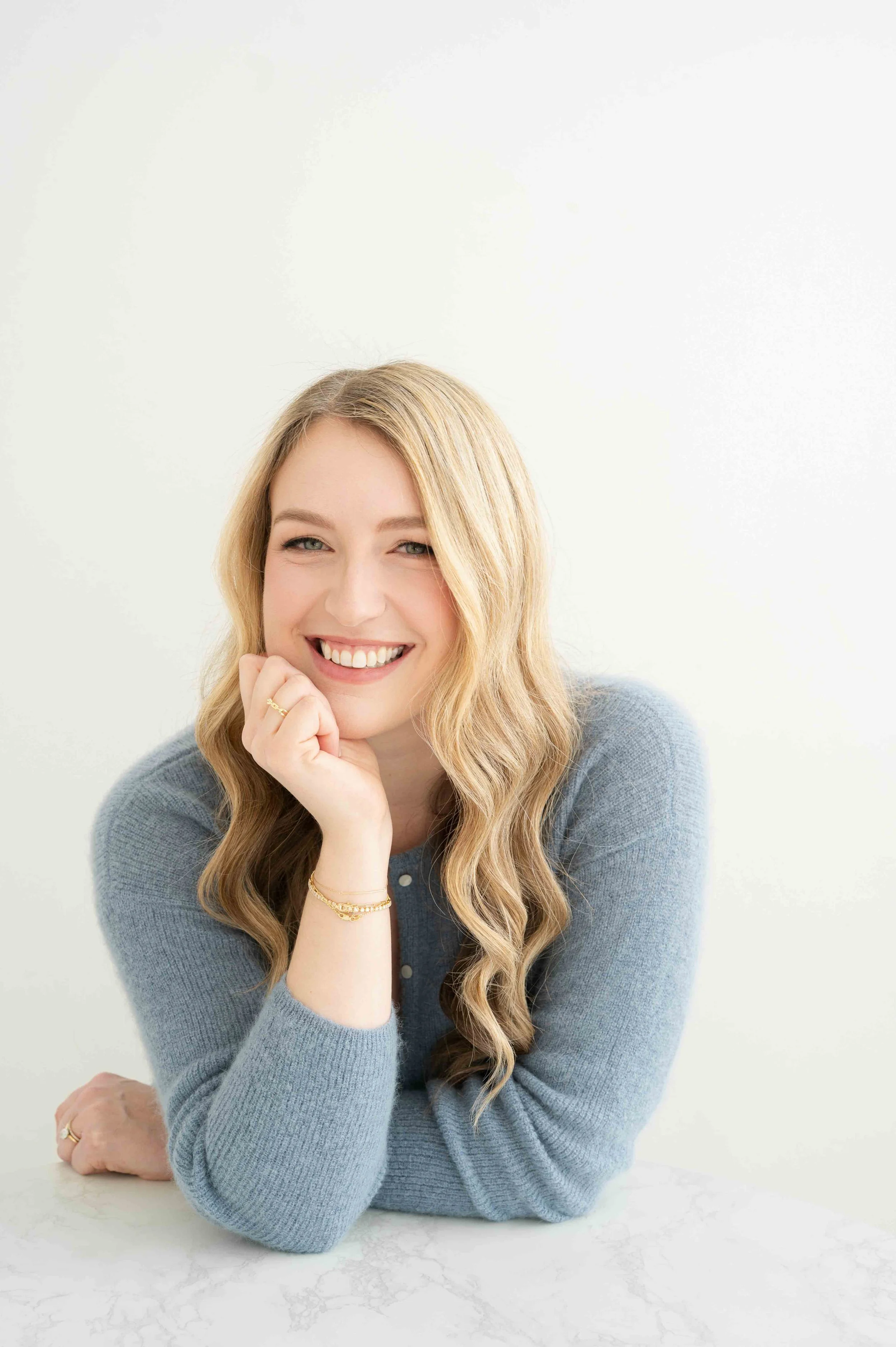 A smiling woman with blonde hair and blue eyes, wearing a blue sweater, rest her chin on her hand, sitting at a table against a bright background.