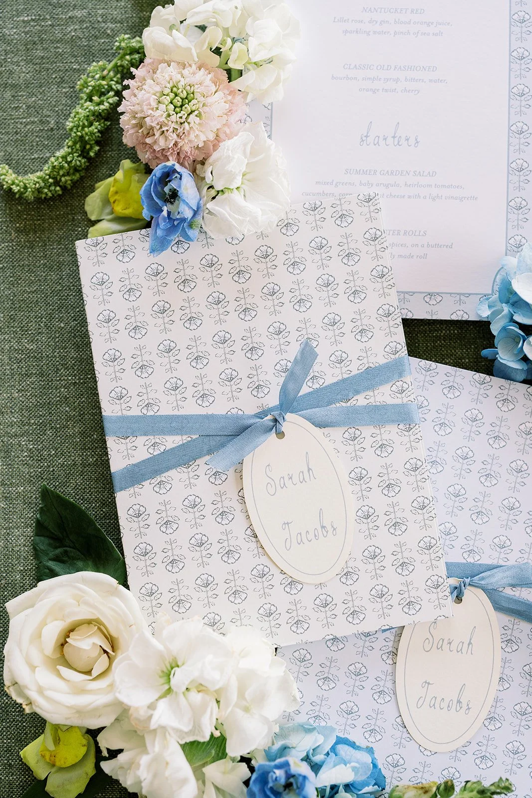 Wedding invitation and place cards decorated with blue ribbons, surrounded by white, pink, and blue flowers on green textured fabric.