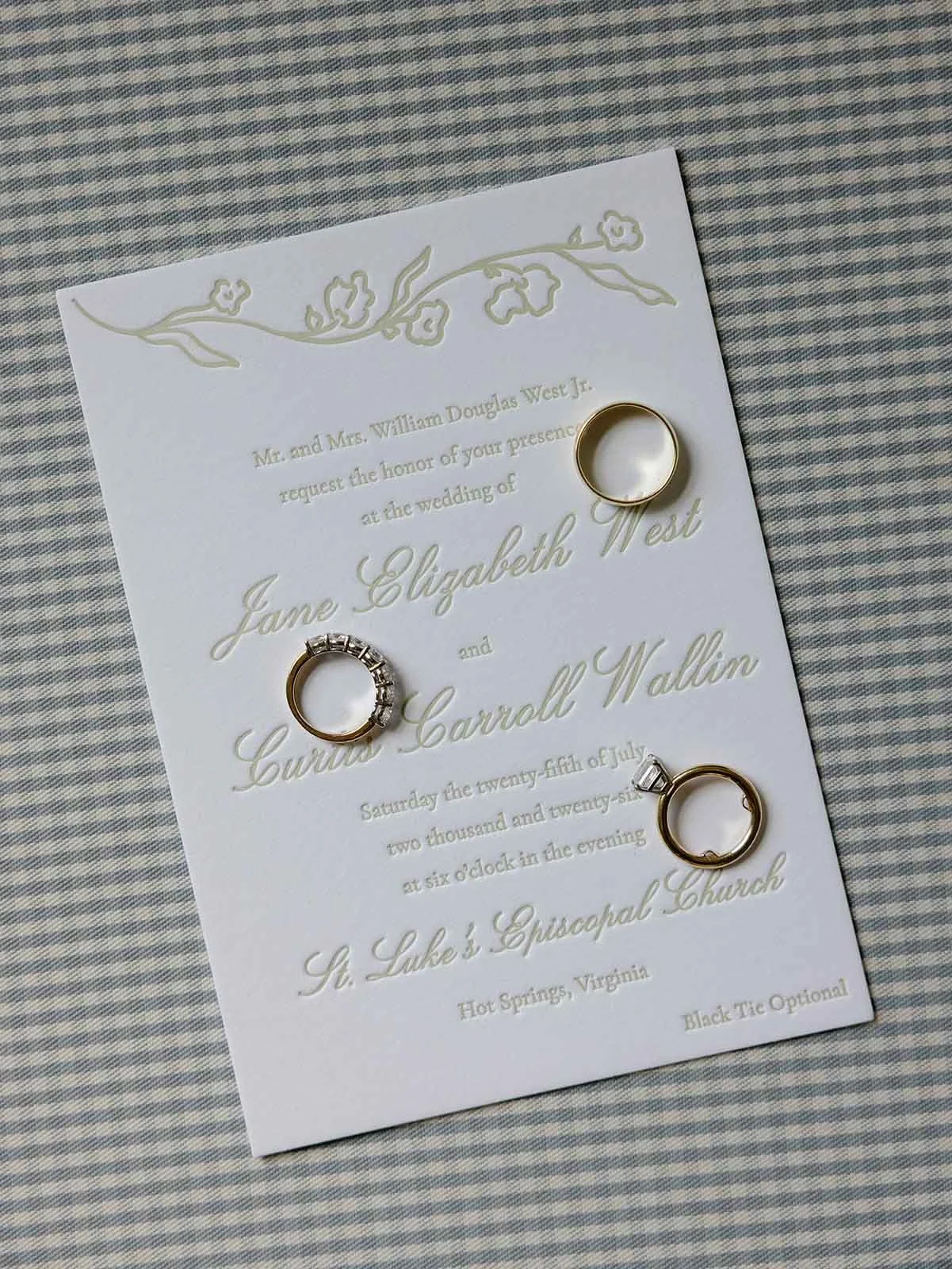 Gingham background behind a wedding invitation with letterpress fonts creating a whimsical wedding invitation and three rings lay on top of the invite