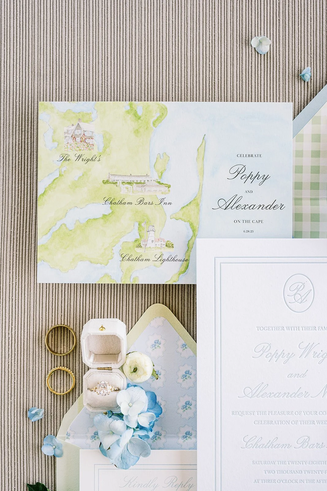 Wedding invitation suite featuring a watercolor map of Cape Cod, a white wedding ring box, rings, and other invitation suite details on a textured brown surface.