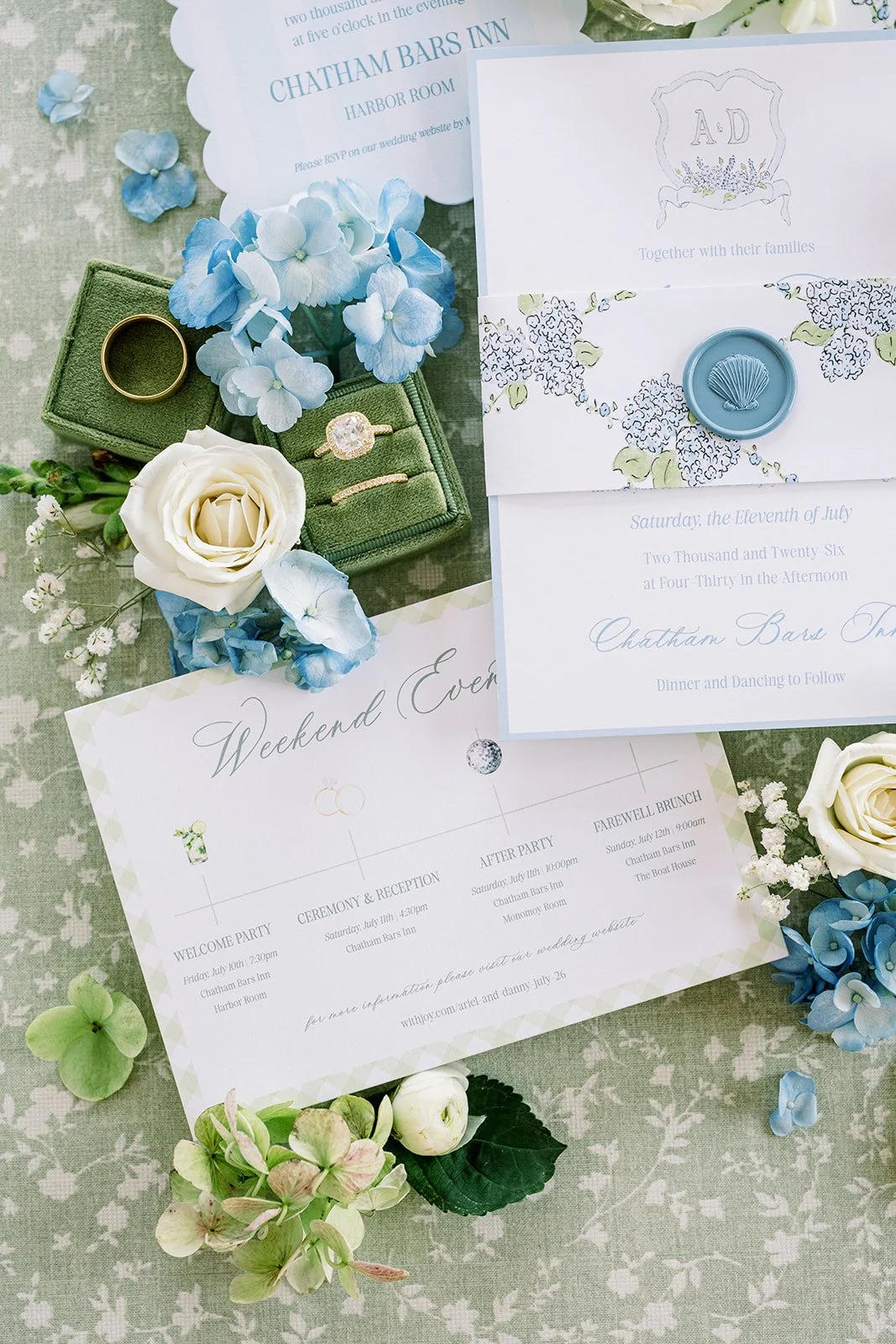 Blue and green weekend events card, invitation, and rehearsal dinner card with fresh florals and a ring box on a green floral background