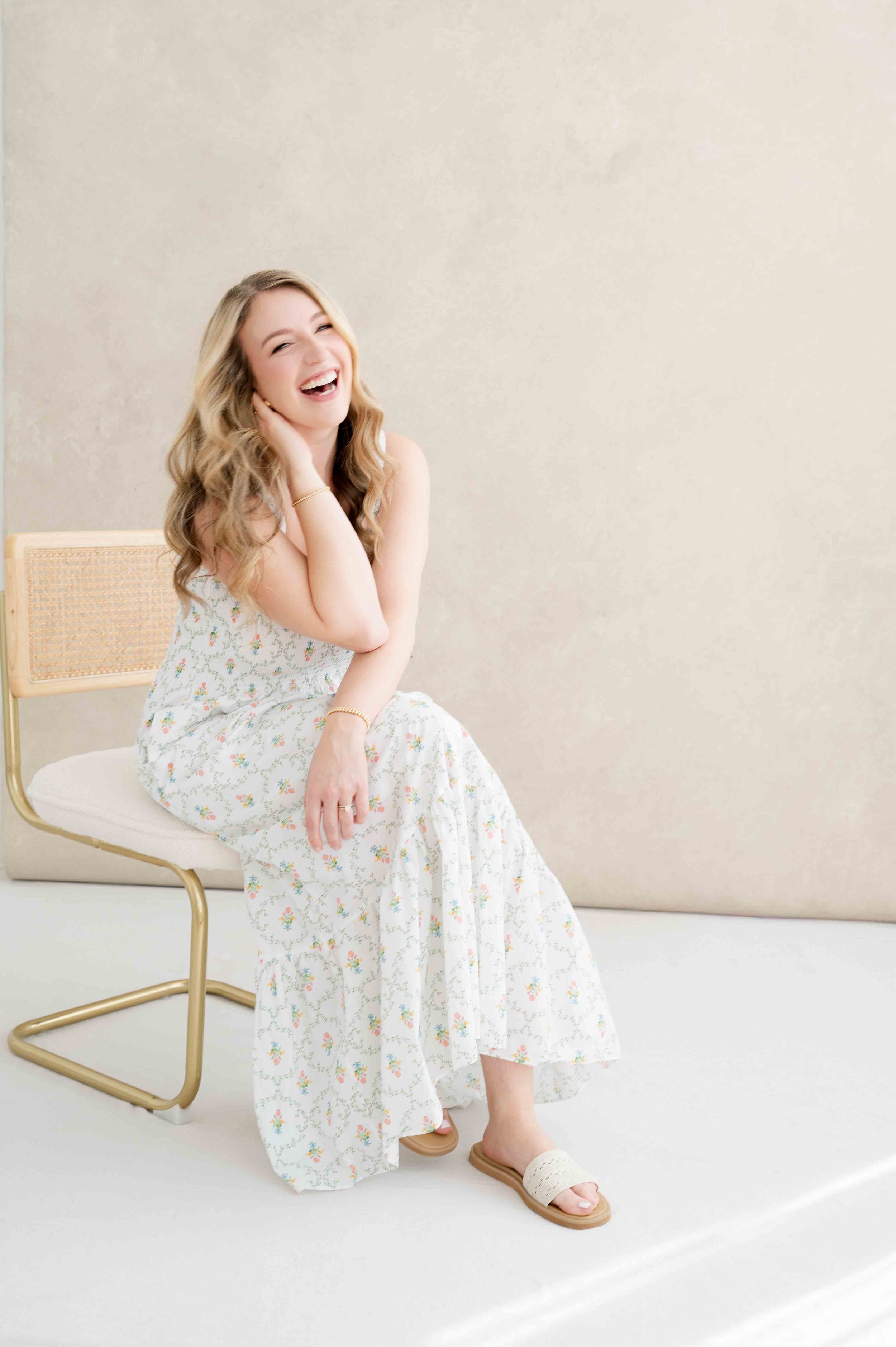 A woman with long wavy hair, wearing a floral dress, sitting on a modern chair with a light-colored background while smiling.