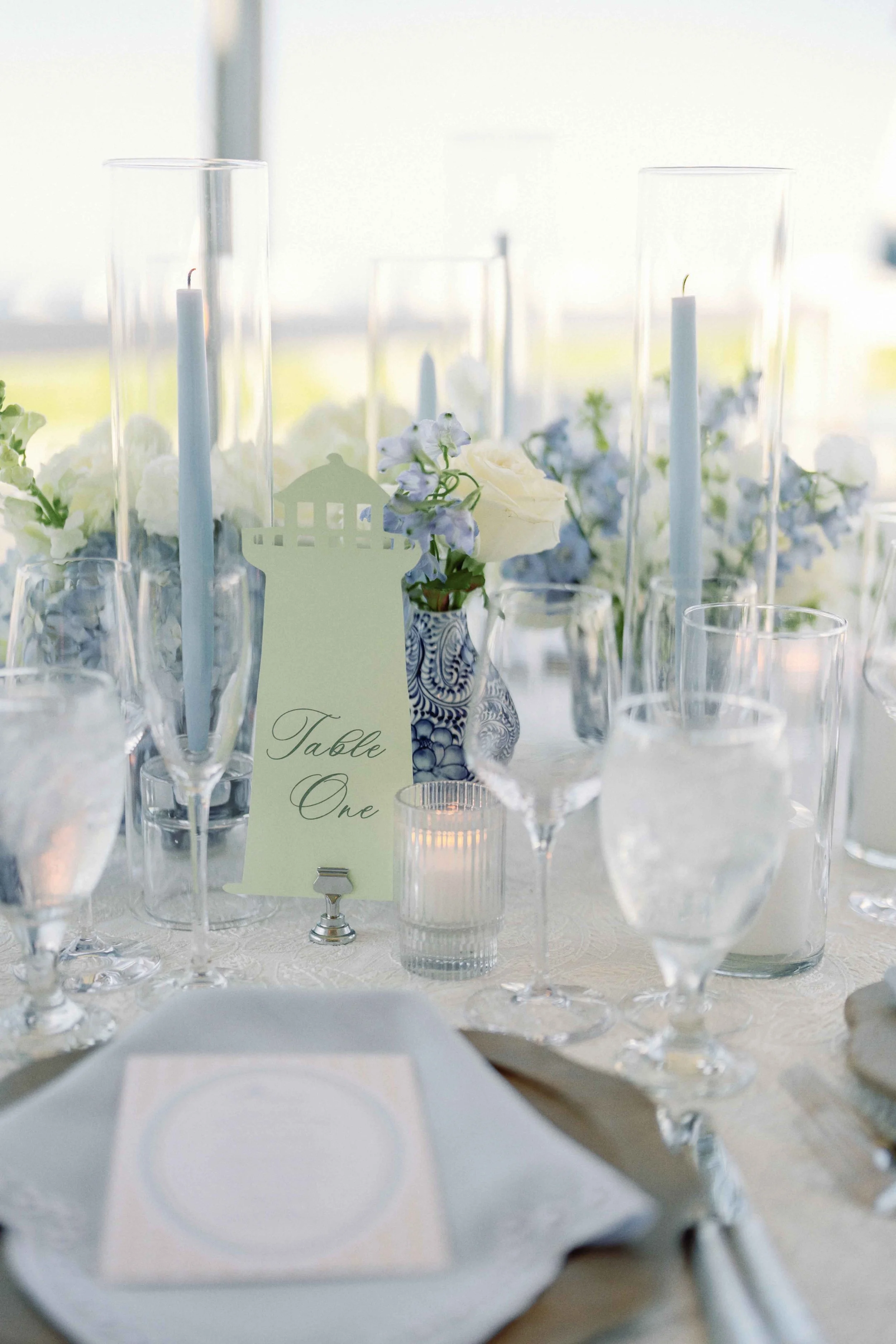 Elegant table setting with tall glass candle holders, flowers, a place card, wine glasses, and a white dinner plate on a white tablecloth over a well-lit background.
