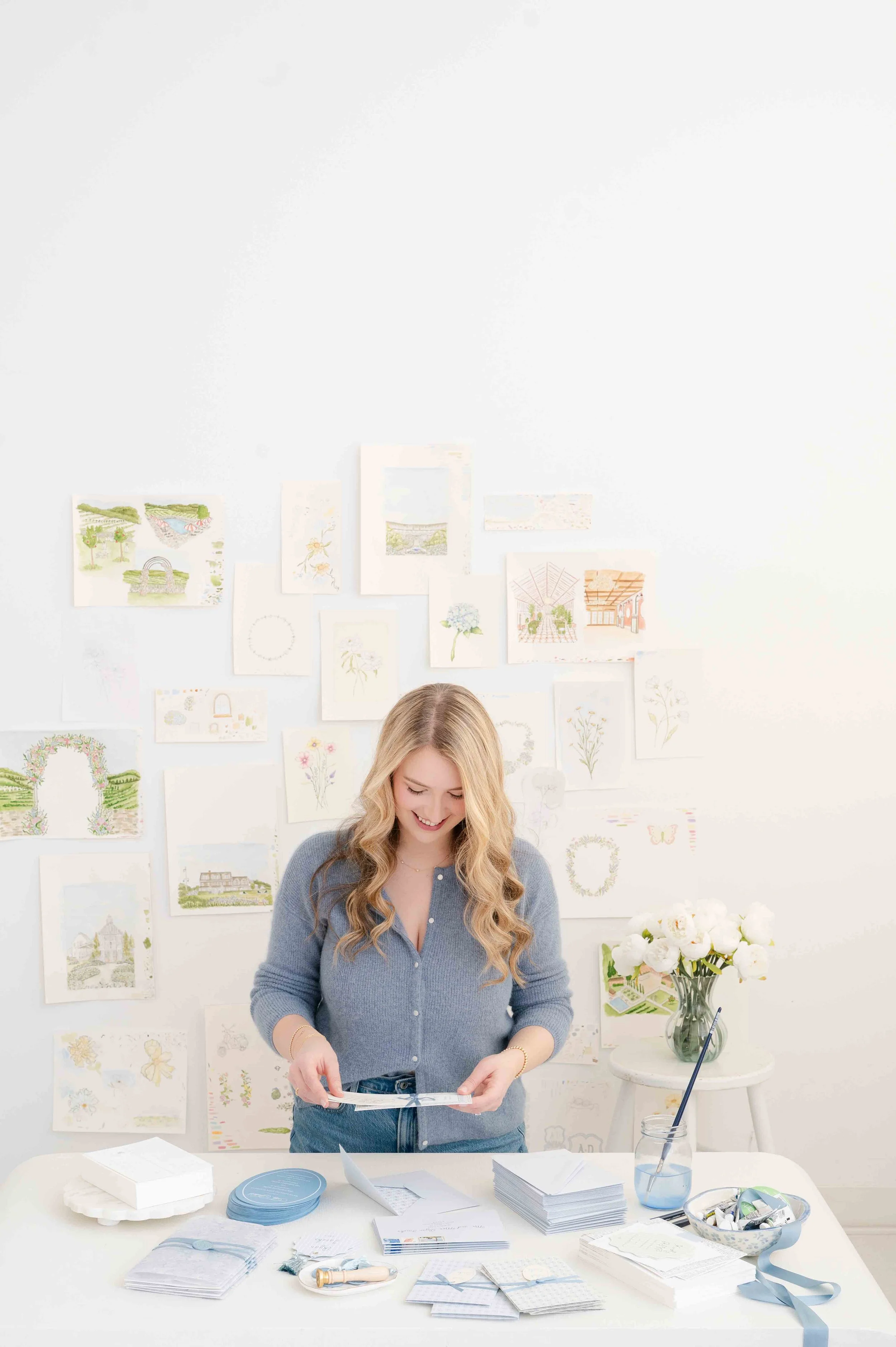 A woman with long blonde hair smiling and looking down at papers she is holding over a white table with craft supplies, with a wall of watercolor sketches and paintings behind her and a vase of white flowers on a small table to her right.