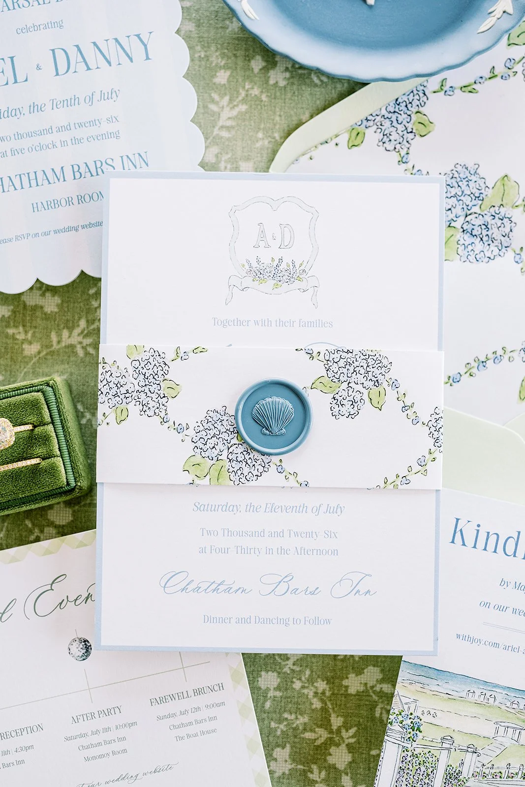 Custom blue, green, and floral wedding stationery on a green floral background with an RSVP card, weekend events card, invitation, envelope, and rehearsal dinner invitation