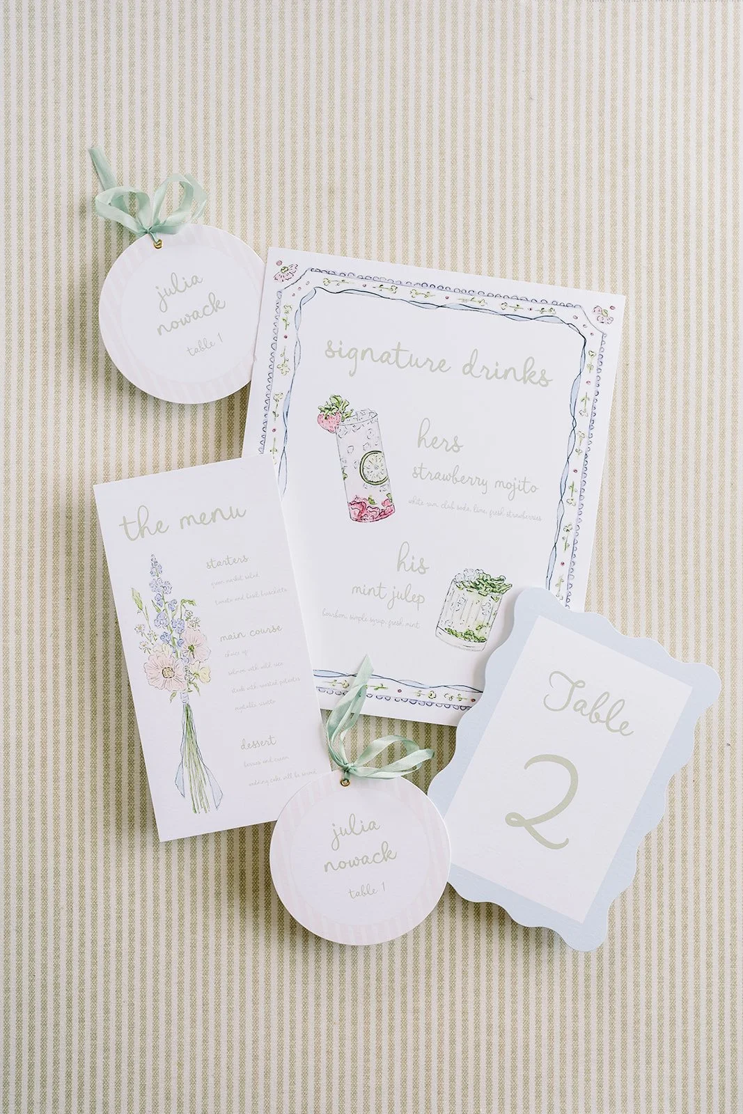 Wedding table settings with signs, a menu, a signature drinks sign featuring a strawberry mojito and mint julep, and name tags, all in light pastel colors on a beige tablecloth.