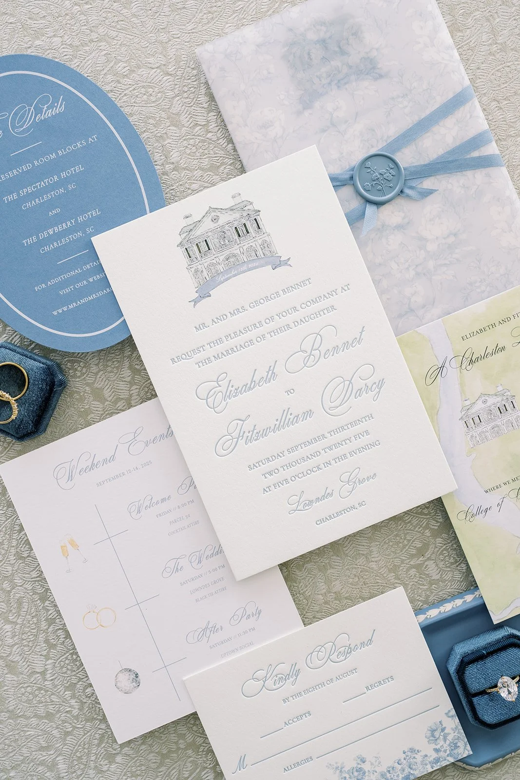 Wedding invitation suite with blue and white theme, including an invitation card, RSVP card, details card, and envelopes, decorated with blue ribbons and wax seals, placed on an elegant textured fabric background.