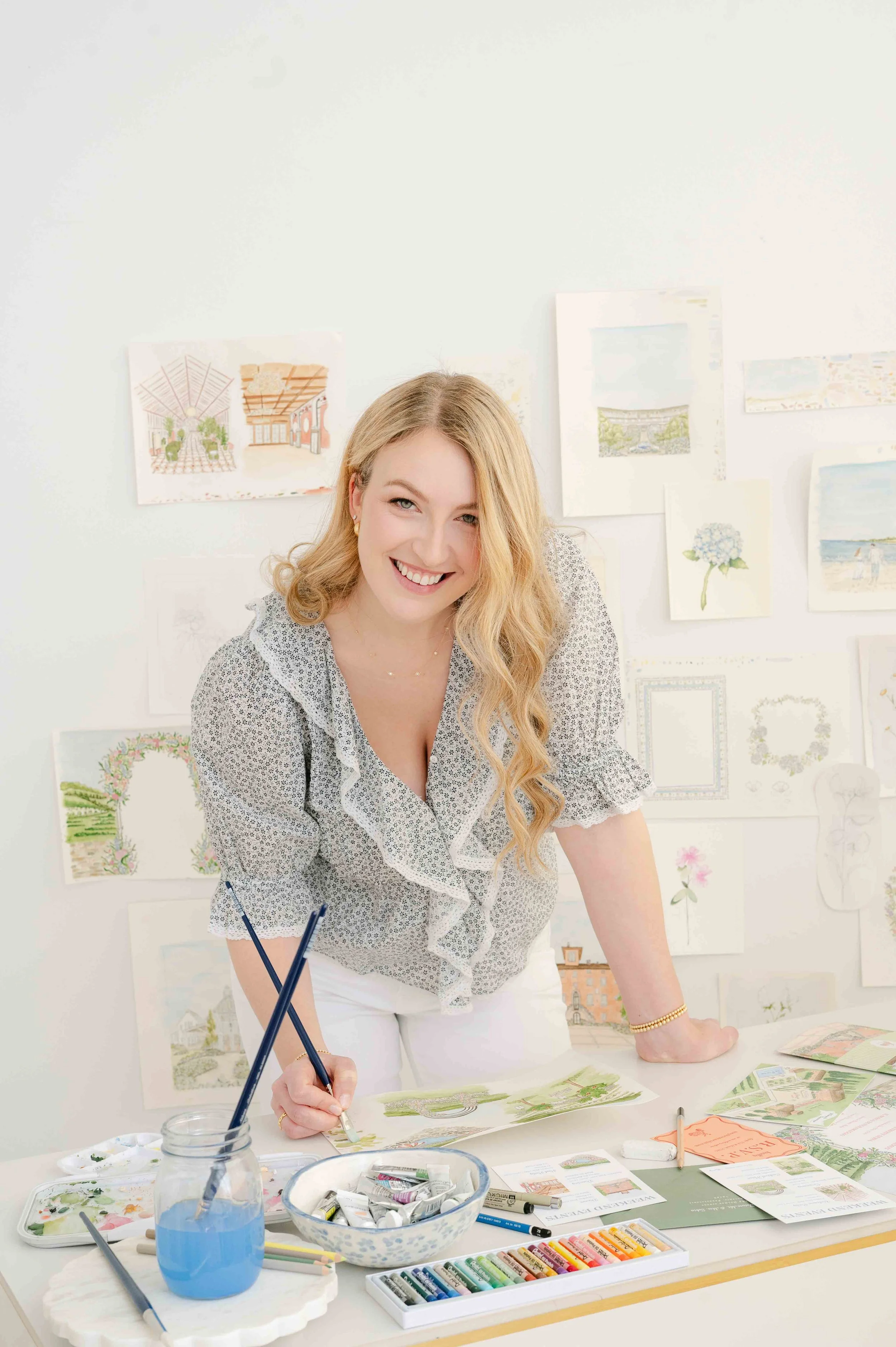 A woman with long blonde hair smiling at the camera, working on watercolor paintings at a table surrounded by art supplies.