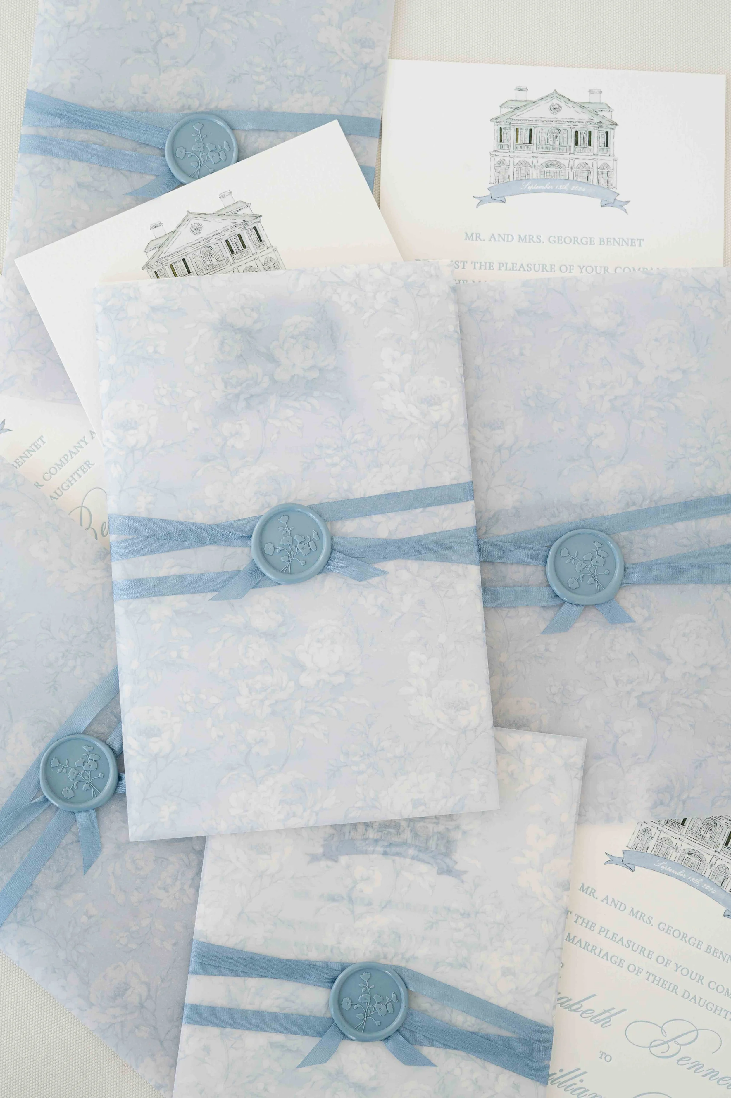 Light blue and white wedding invitations with floral patterns, tied with matching blue ribbons and wax seal stamps featuring a floral design.