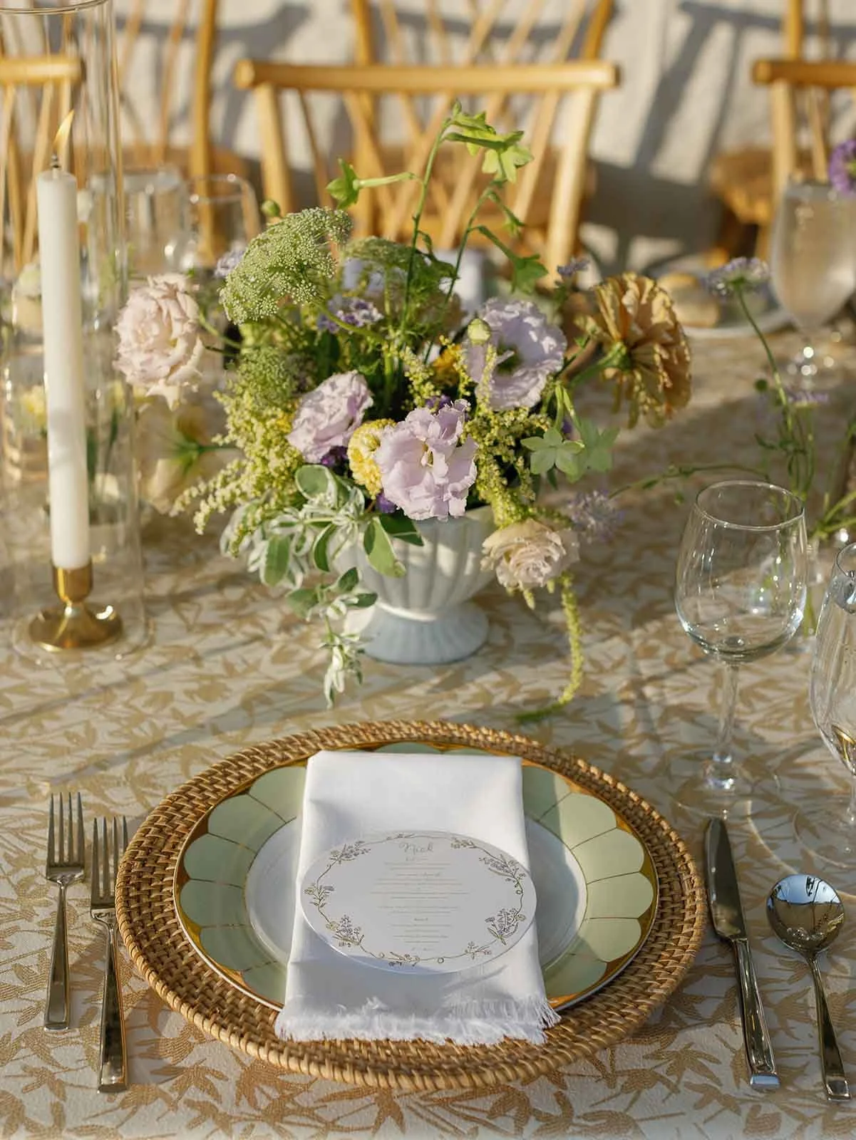 Outdoor wedding tablescape featuring a plate, placemat, napkin, and circle menu on top of a table featuring florals, silverware, and candles