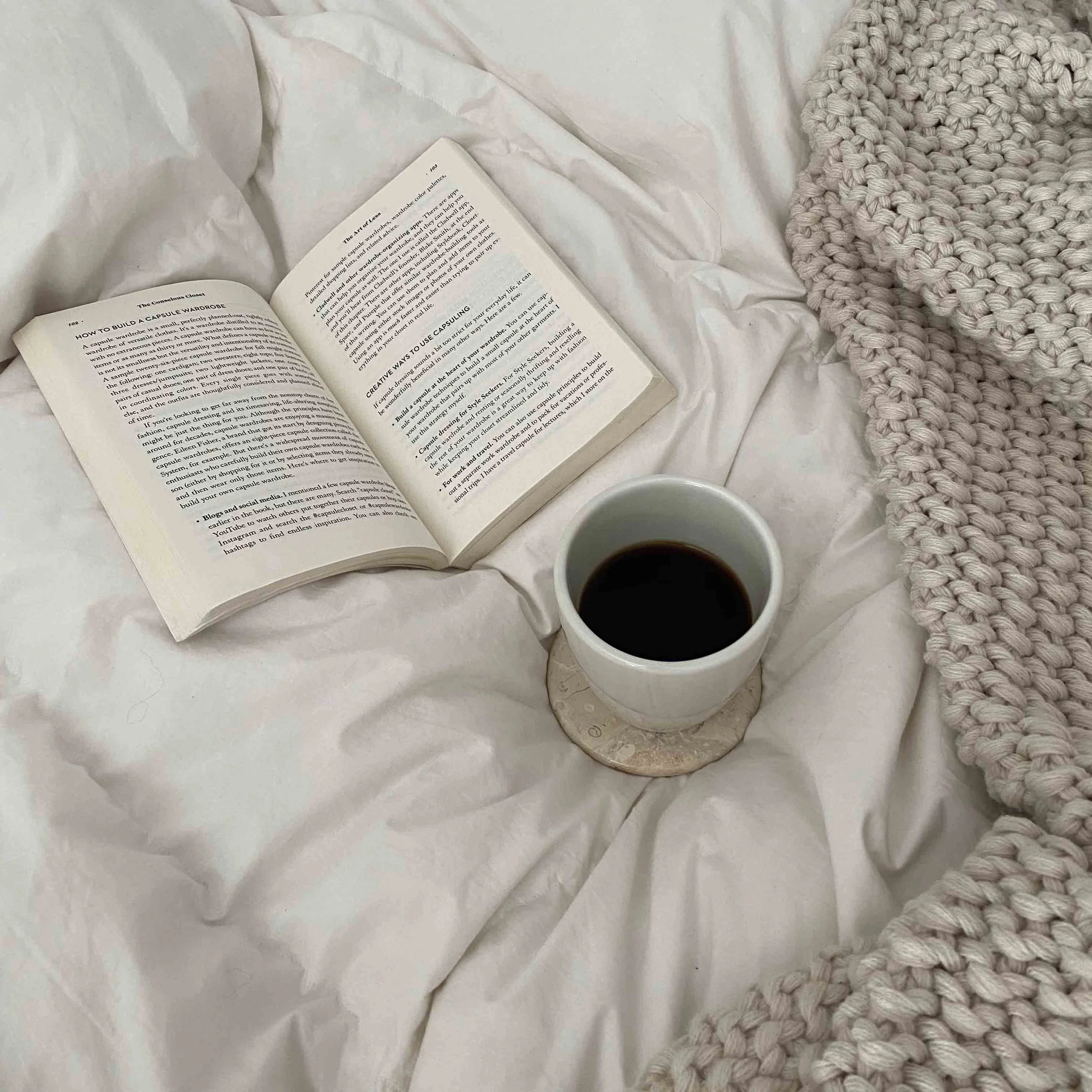 Open book on white bedsheet next to a white mug of coffee and a chunky tan blanket.
