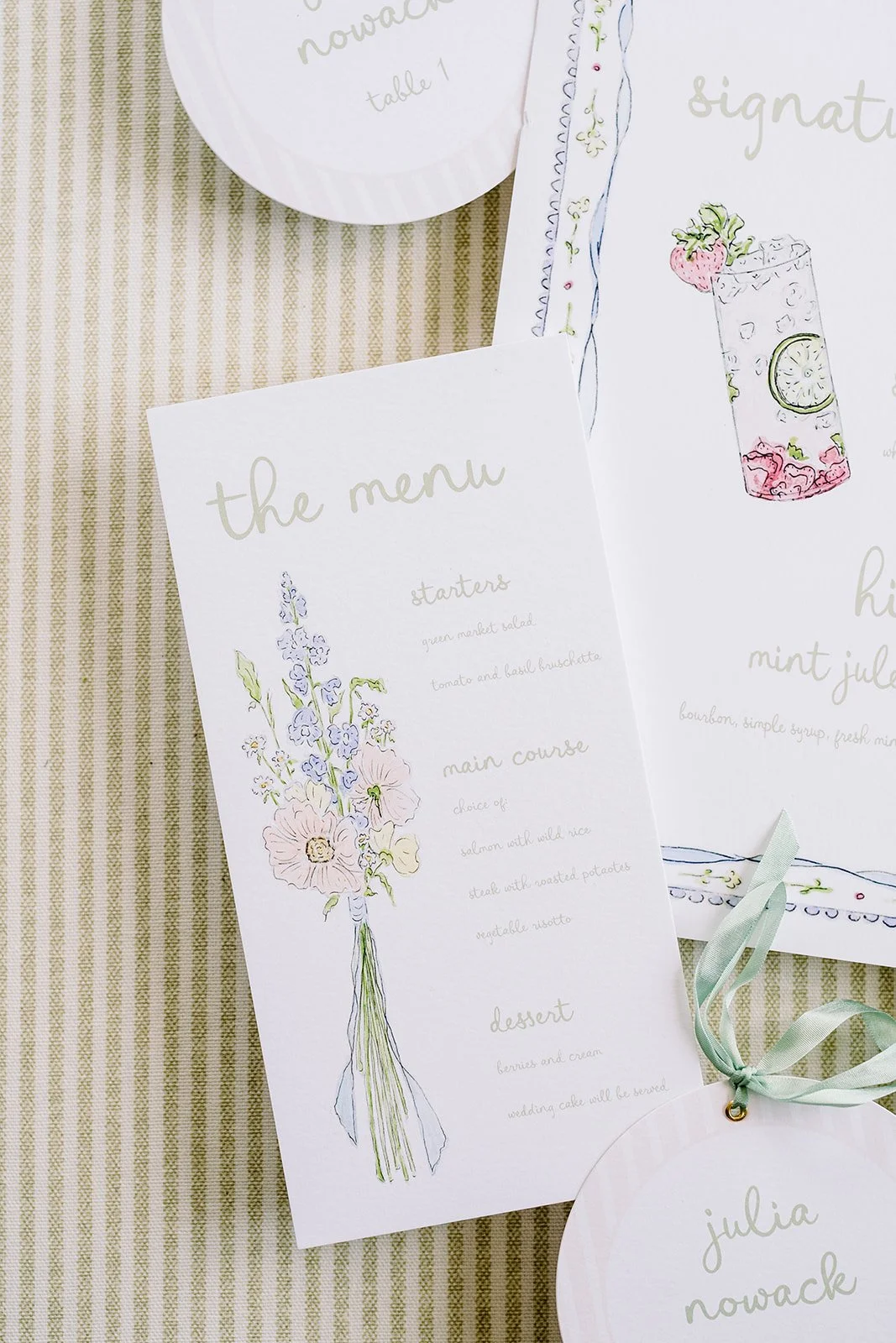 Wedding or event menu printed on white paper with pastel watercolor illustrations of flowers, placed on a green and white striped tablecloth, with a name tag and other wedding stationery.