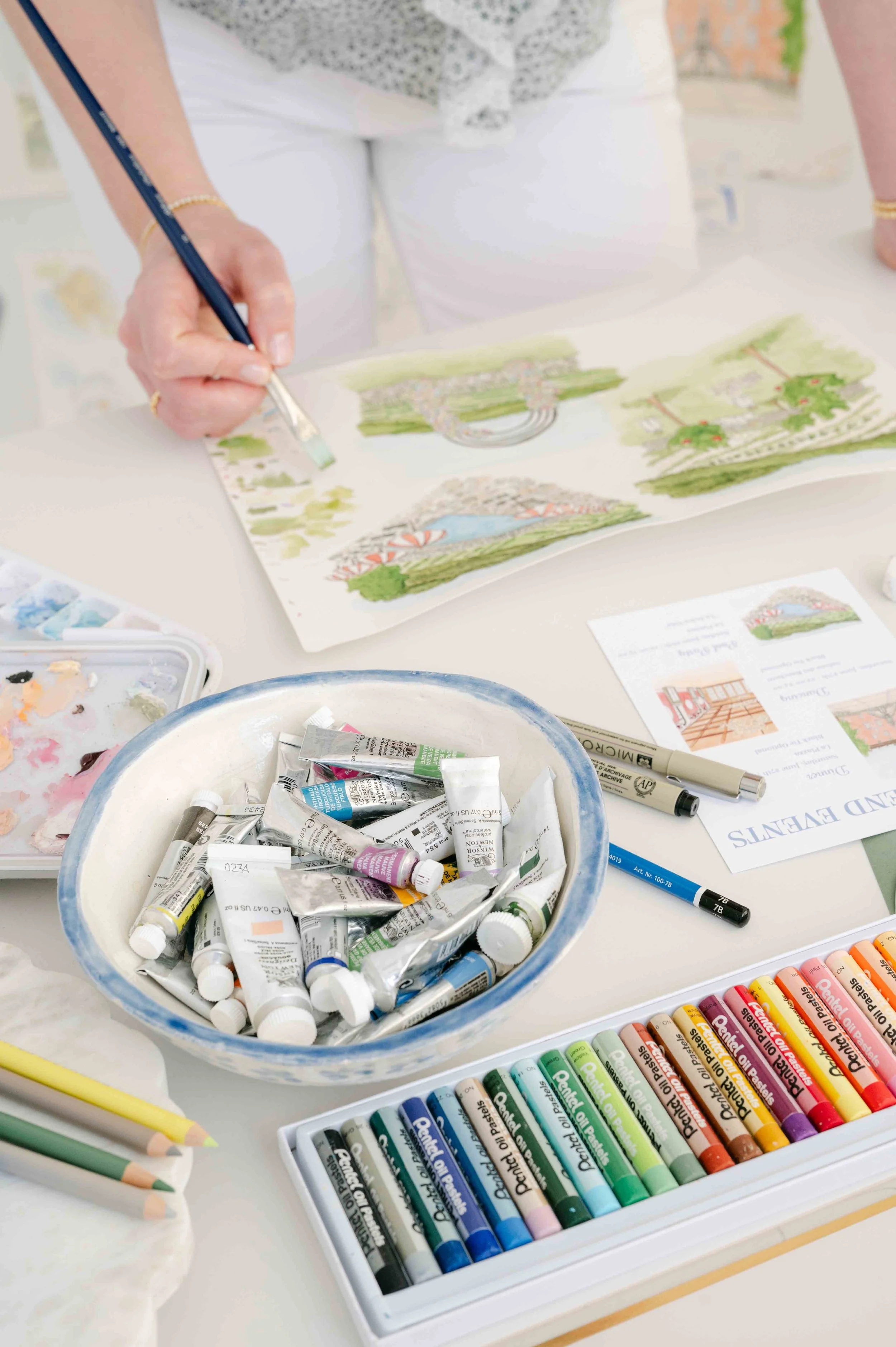 An artist paints landscape illustrations with watercolor paints and colored pencils on a white table.