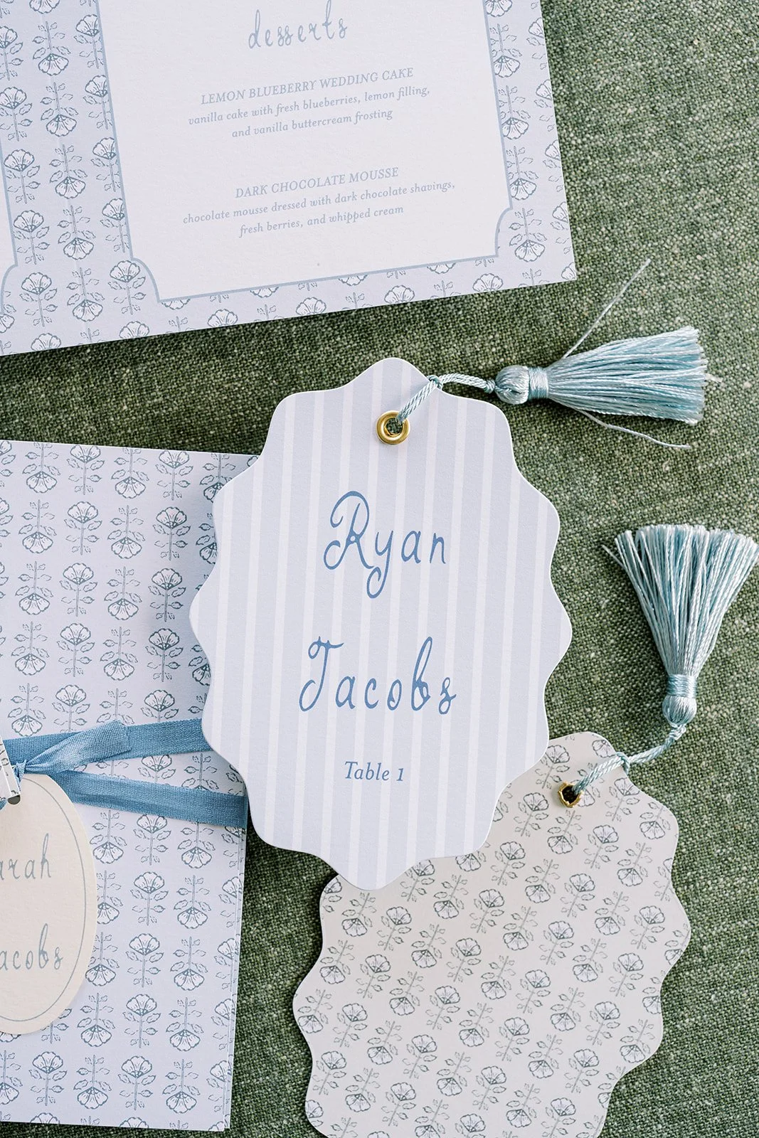 Wedding reception seating card with the names and table numbers, decorated with light blue tassels, surrounded by matching paper decorations and a menu card.