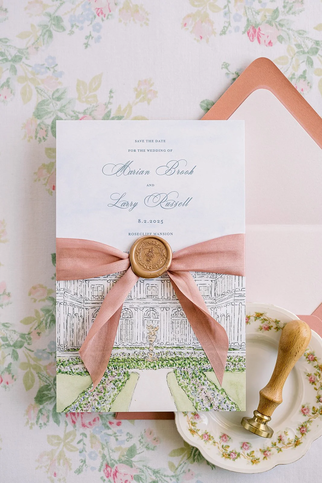 A wedding invitation card decorated with a pink ribbon and wax seal, featuring an illustrated watercolor building and floral details, placed on a floral tablecloth with a pink envelope and a decorative stamping wax seal stamp.