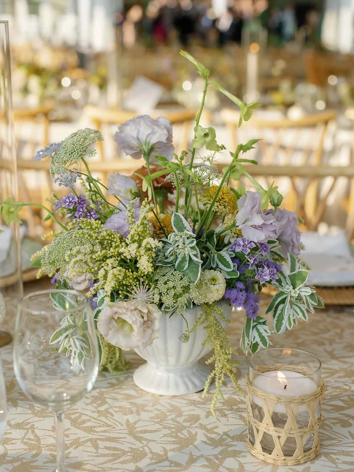 Beautiful green, yellow, and purple flowers in a white vase surrounded by candles and drinkware at a whimsical outdoor wedding 