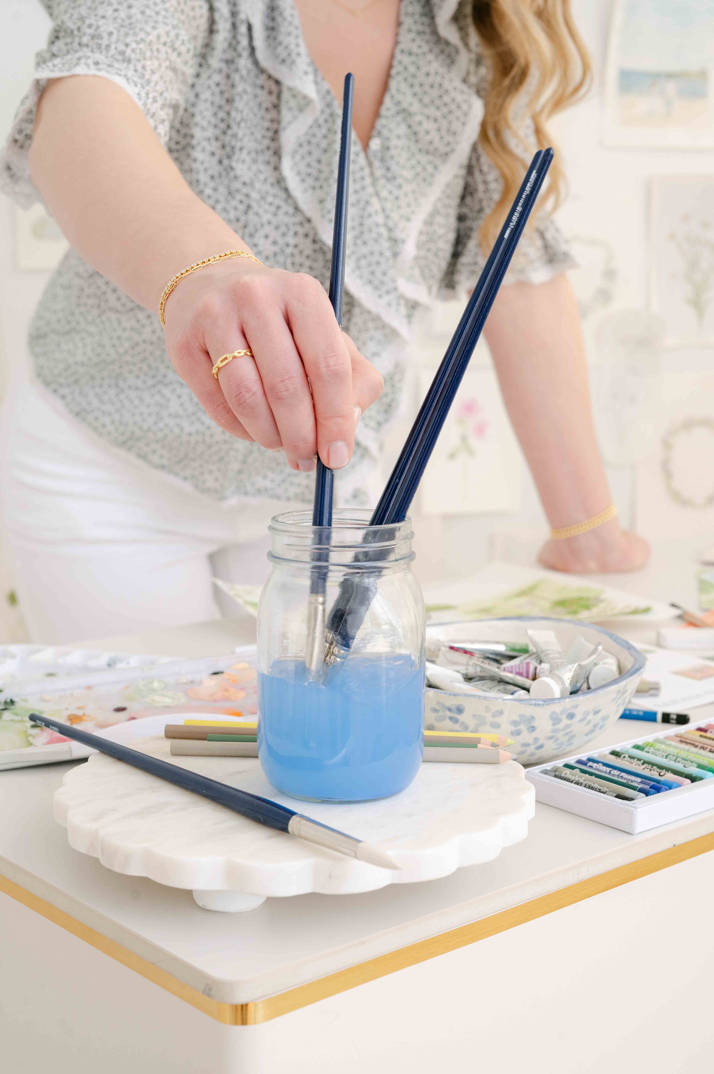 A woman arranging paintbrushes in a jar filled with blue water, with art supplies like paints and colored pencils on a table.