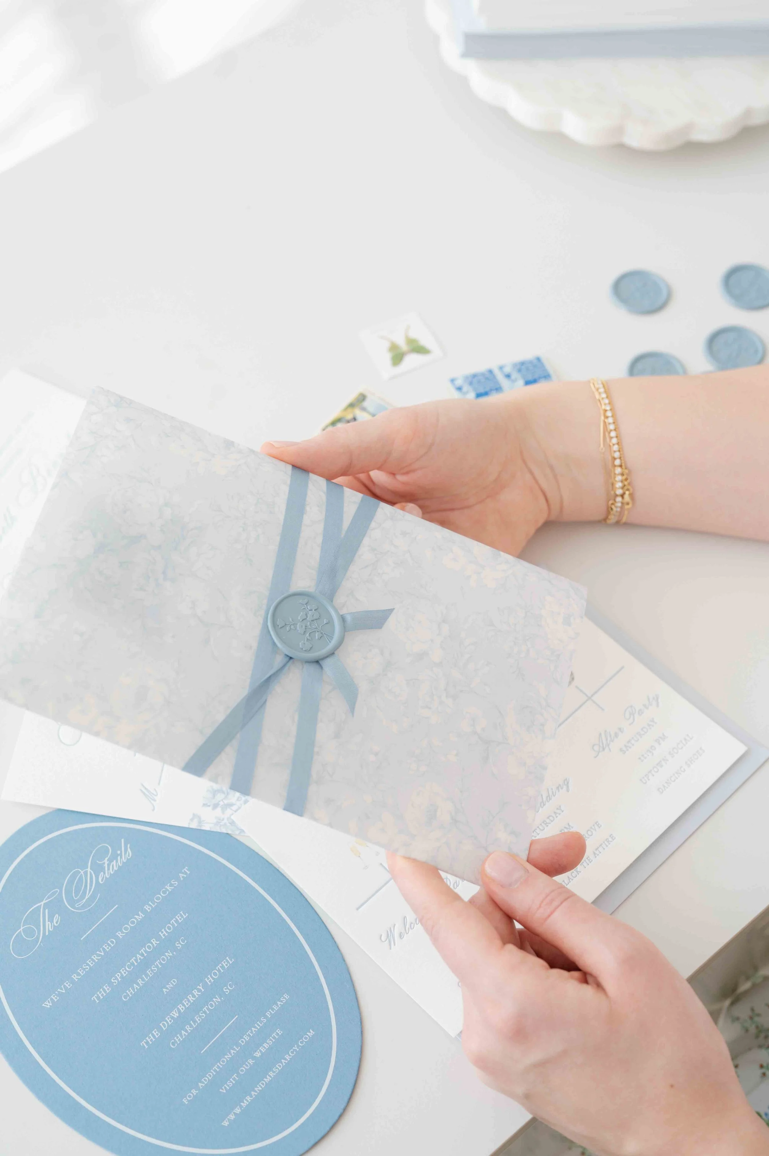 A person holding a wedding invitation with a blue ribbon and wax seal. On the table are wedding stationery details, wax seals, and more.